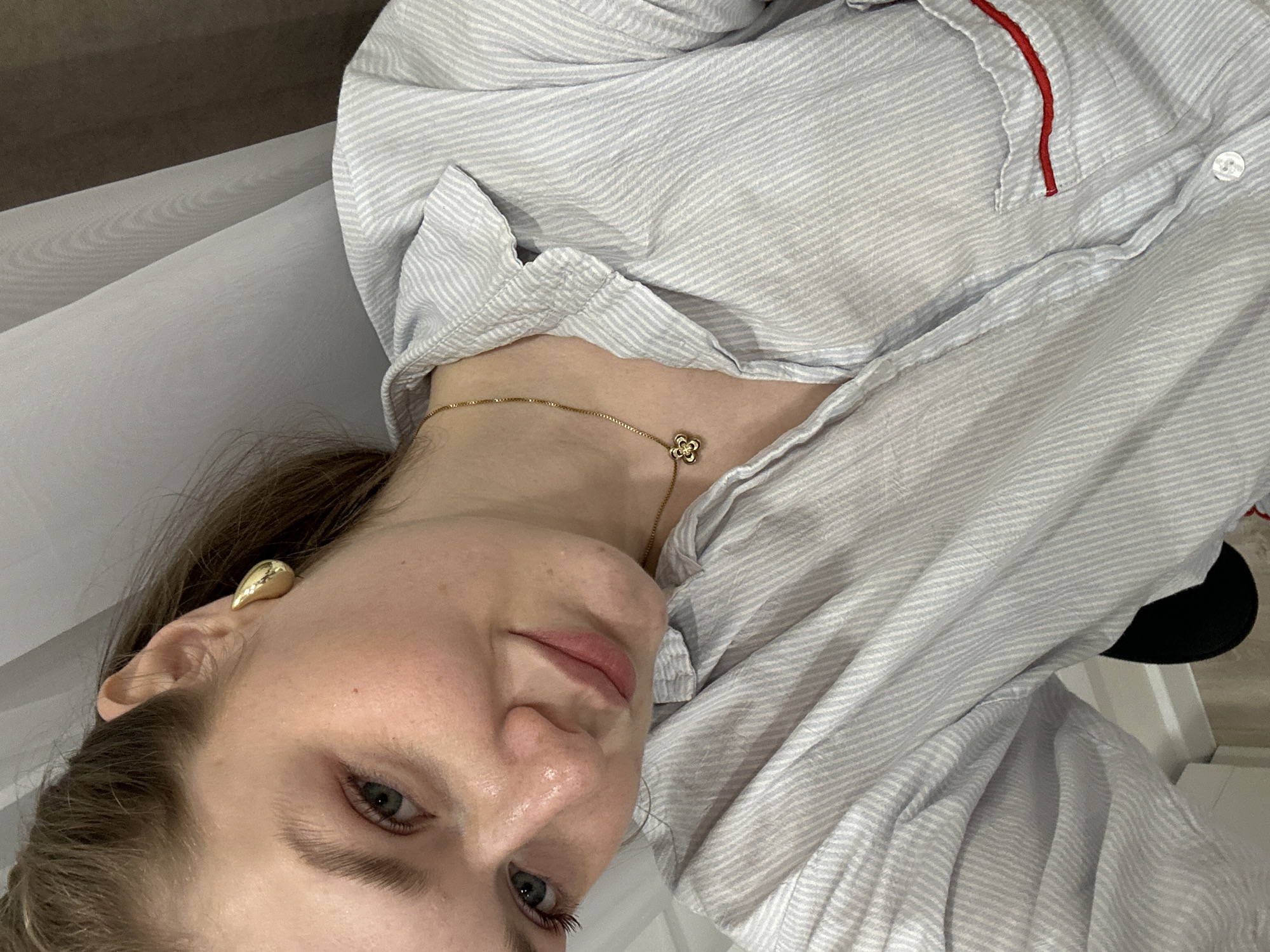 A woman is pictured indoors in a close-up shot. She is wearing a light blue and white striped button-down shirt, and a gold necklace with a flower-shaped pendant is visible. She also has gold teardrop earrings. No information is available to place the location in the city of Kazan, Russia.