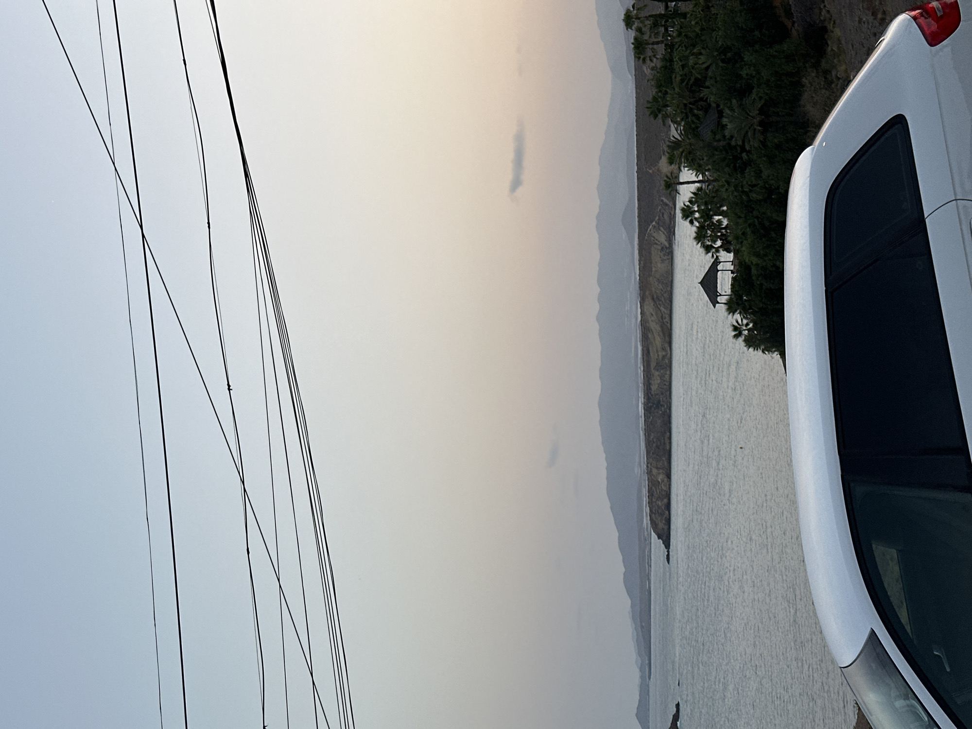 In Bolnuevo, Spain, a white car is parked on a hillside overlooking the sea. The car's rear is in the foreground, with its roof and rear window visible. Beyond the car, the calm sea stretches out, meeting a distant, hazy coastline with rolling hills. A small gazebo-like structure is nestled amongst lush green trees and palm trees on the slopes leading down to the water. Power lines are strung across the top of the image, silhouetted against a pale, twilight sky. The overall scene suggests a peaceful, coastal setting at dusk.