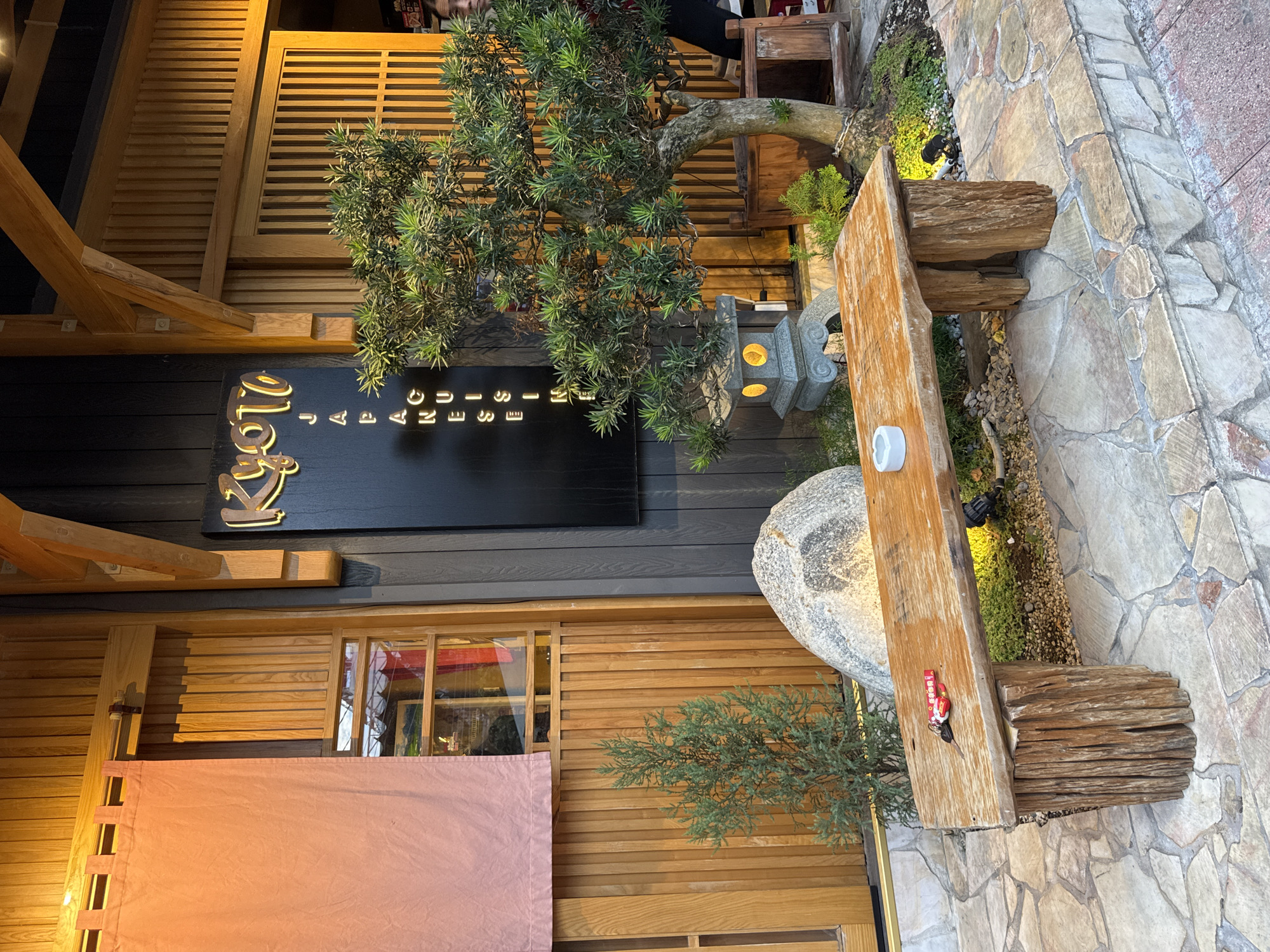 A rustic wooden bench with two people's belongings, including keys and a red object, sits on a stone patio outside a Japanese restaurant. The restaurant features a traditional wooden facade with a pink curtain over a window. A sign above the entrance reads "Kyoto Japanese Cuisine." A small, stylized Japanese lantern and a potted tree are also visible. The scene is lit by warm, ambient lighting.