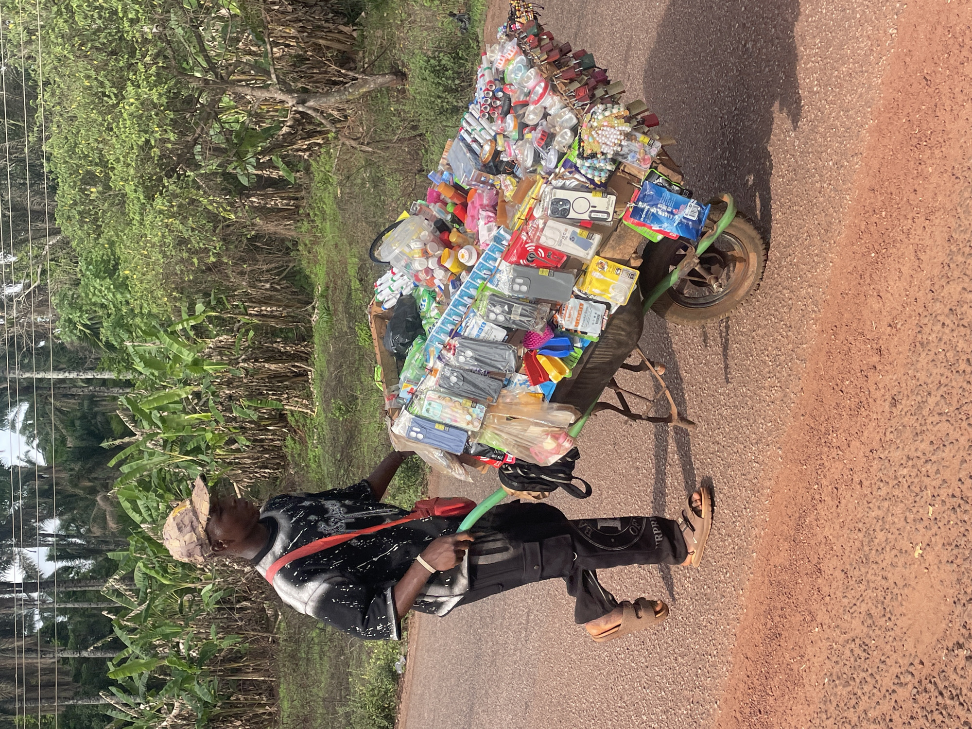 A man walks along a paved road in Ogbe, Nigeria, pulling a heavily laden, single-wheeled cart. The cart is filled with a variety of goods, including phone cases, locks, and plastic containers. The man is wearing a patterned shirt, black pants, sandals, and a hat. He is pulling the cart with a green handle. The road is surrounded by vegetation.