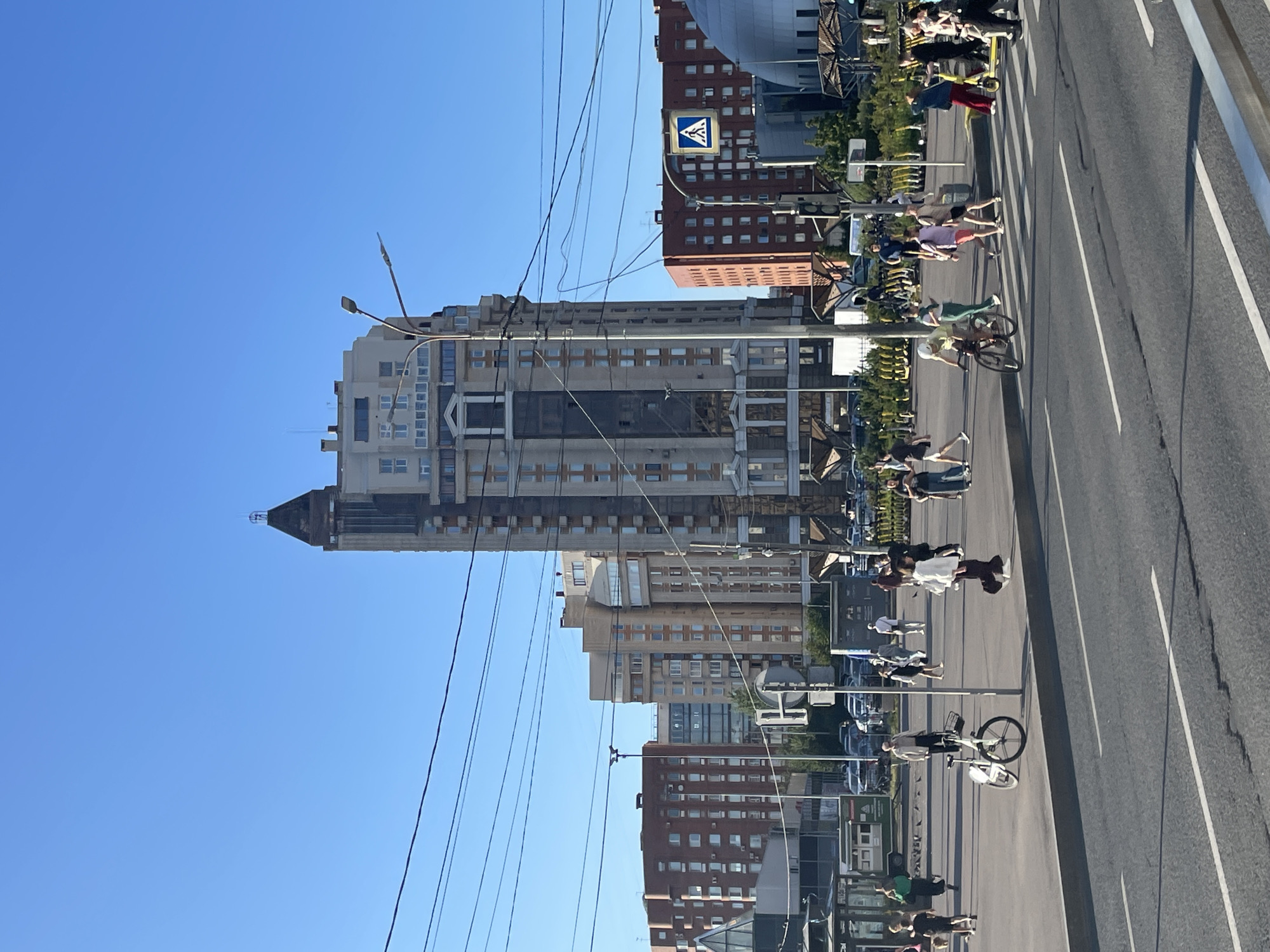 The image depicts a street scene in Ladozhskii Trudposelok, Russia, on a clear, sunny day. In the background, a prominent, tall, modern skyscraper with a dark, pointed roof dominates the skyline, flanked by several mid-rise apartment buildings, some brick-red and others cream-colored. In the middle ground, a pedestrian plaza is active with people walking and several bicycles. One bicycle is stationary near a lamppost. A crosswalk is visible in the foreground with pedestrians crossing. There are also streetlights and overhead power lines stretching across the sky. The scene appears to be a busy urban environment during daylight hours.