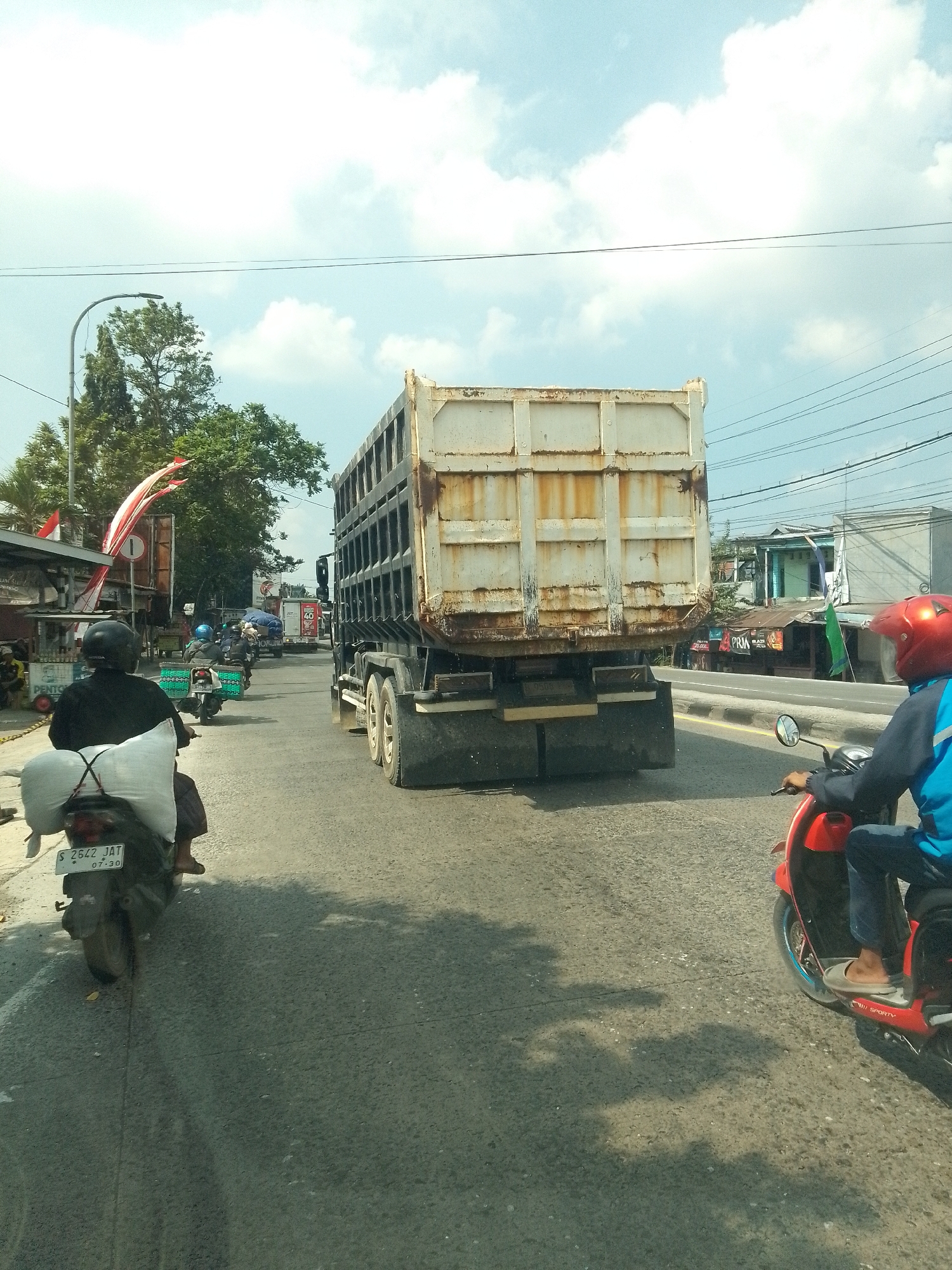 In Bedahan, Indonesia, on a sunny day with scattered clouds, a busy street scene unfolds. A large, white dump truck with a weathered exterior dominates the right side of the frame, its rear wheels and chassis clearly visible as it travels on the asphalt road. Ahead of the truck, a red scooter rider, wearing a blue jacket and a red helmet, navigates the traffic. To the left of the truck, a motorcycle, with a rider wearing a black helmet and carrying a large white bundle secured to the back, is also in motion. Further ahead on the left, another motorcycle with two riders, both wearing helmets, is visible, and beyond that, a lighter-colored truck with a red sign displaying "60" can be seen. The surrounding environment includes trees, streetlights, overhead wires, and various buildings and shops lining the road, one of which has a red and white Indonesian flag prominently displayed. The road surface shows signs of wear and tear, with tire marks and a yellow dividing line. Shadows of vehicles and trees are cast on the asphalt.