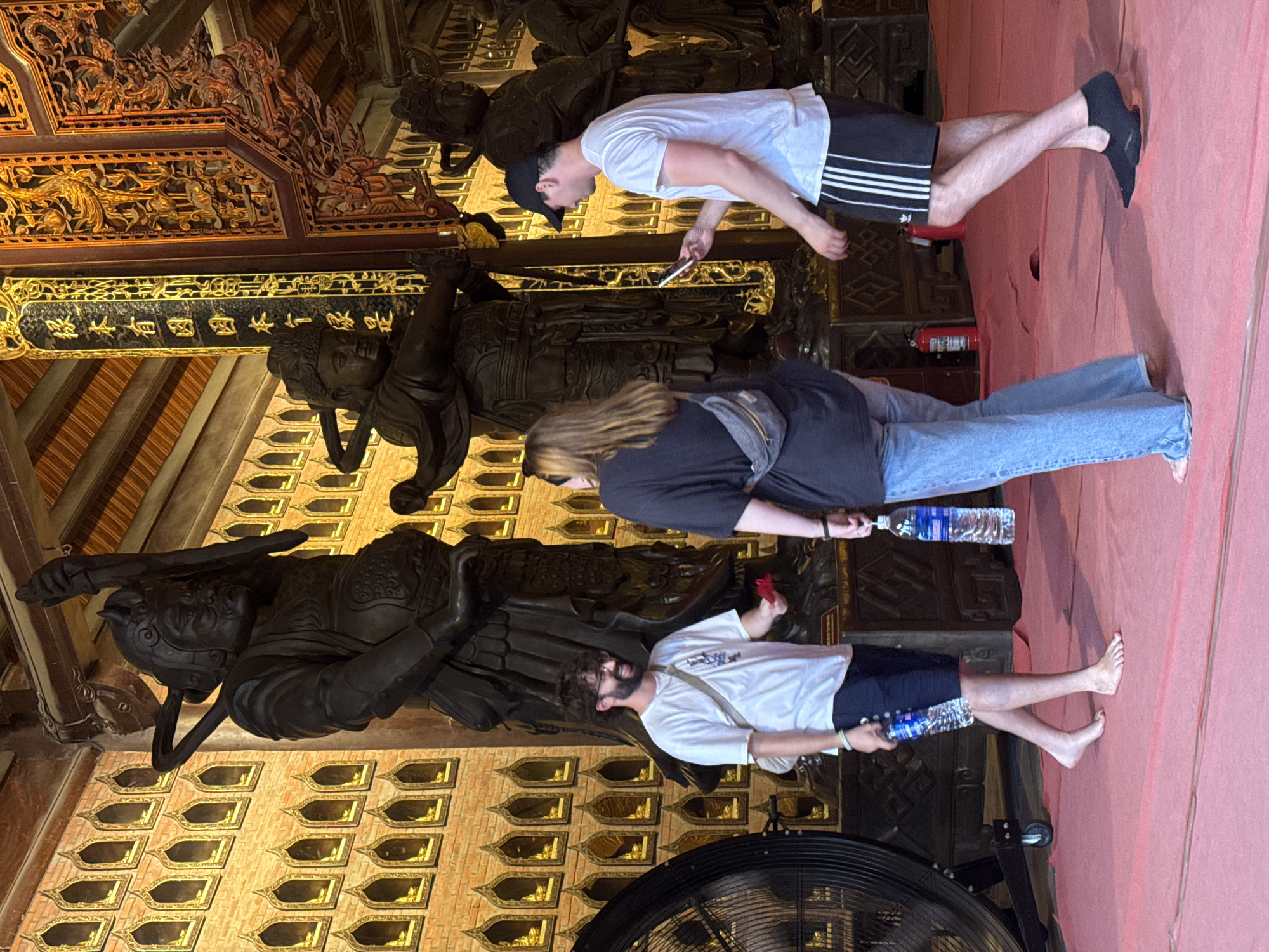 In the city of Tây Hoa Lư Ward, Vietnam, three individuals are present inside a temple with ornate wooden carvings and numerous small niches containing golden objects on a brick wall. In the foreground, a man with a beard and glasses, wearing a white t-shirt and blue shorts, stands barefoot and holds a red object and a water bottle. To his right, a woman with blonde hair, wearing a black t-shirt and blue jeans with a fanny pack, also stands barefoot and holds a water bottle. On the far right, a man wearing a white t-shirt, black shorts with white stripes, and a black baseball cap is looking at his phone. He is wearing black socks with a slip-on design. Two large warrior statues are visible in the background. The floor is covered with a red carpet, and a large black fan is positioned on the left side of the frame. A red fire extinguisher is visible behind the woman.