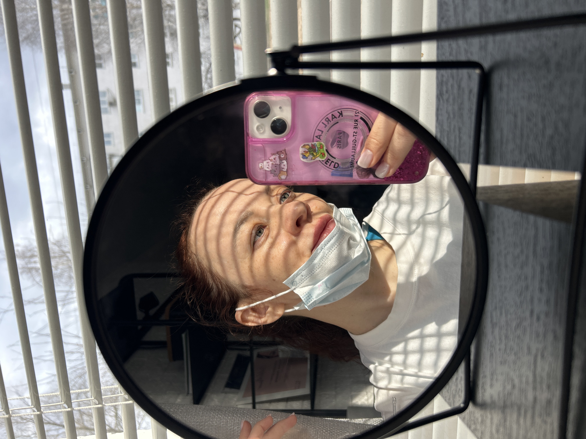 A person wearing a light blue disposable face mask and a white t-shirt is reflected in a round mirror. The person is holding a pink smartphone with stickers on its case, taking the selfie. The background includes a window with closed blinds, and the presence of buildings outside suggests an urban environment. The city of Chaikovsky, Russia is not visibly evident in the photograph.