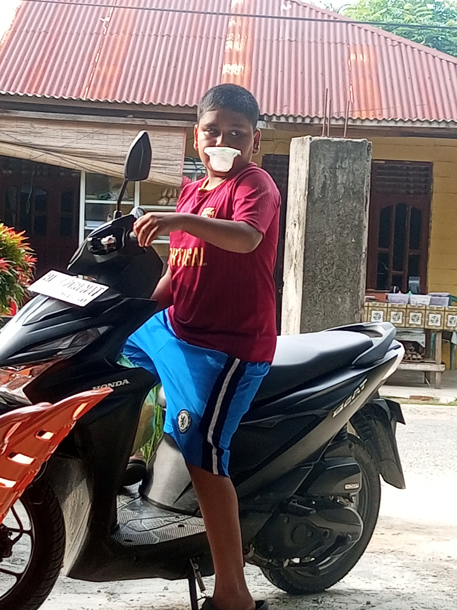 A young boy is seated on a black and orange Honda Beat scooter, holding a white cup to his mouth. He is wearing a red t-shirt with white text that reads "ORTICAL" and blue shorts with white stripes. The scooter is parked on a dirt or gravel surface in front of a house with a red corrugated metal roof. A concrete pillar stands to the boy's right, and a table with patterned cloth and containers is visible behind it. The scene appears to be outdoors in a residential area.