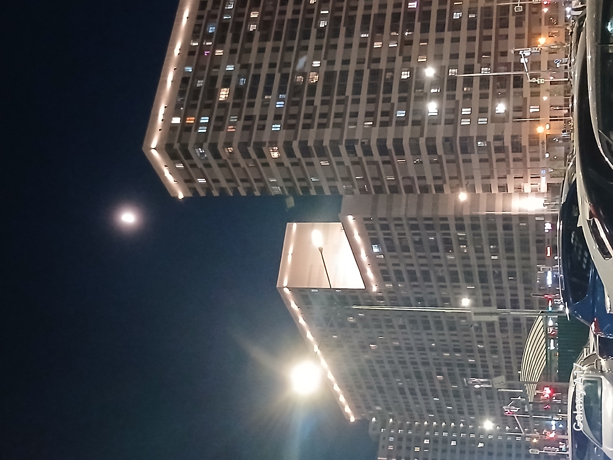The image shows a nighttime scene with a building illuminated by numerous windows and street lights in Yekaterinburg, Russia. The sky is dark blue with a bright moon visible in the upper-left. In the lower half, there is the reflection of the building along with the glare from a streetlight. There are also cars parked along the street.