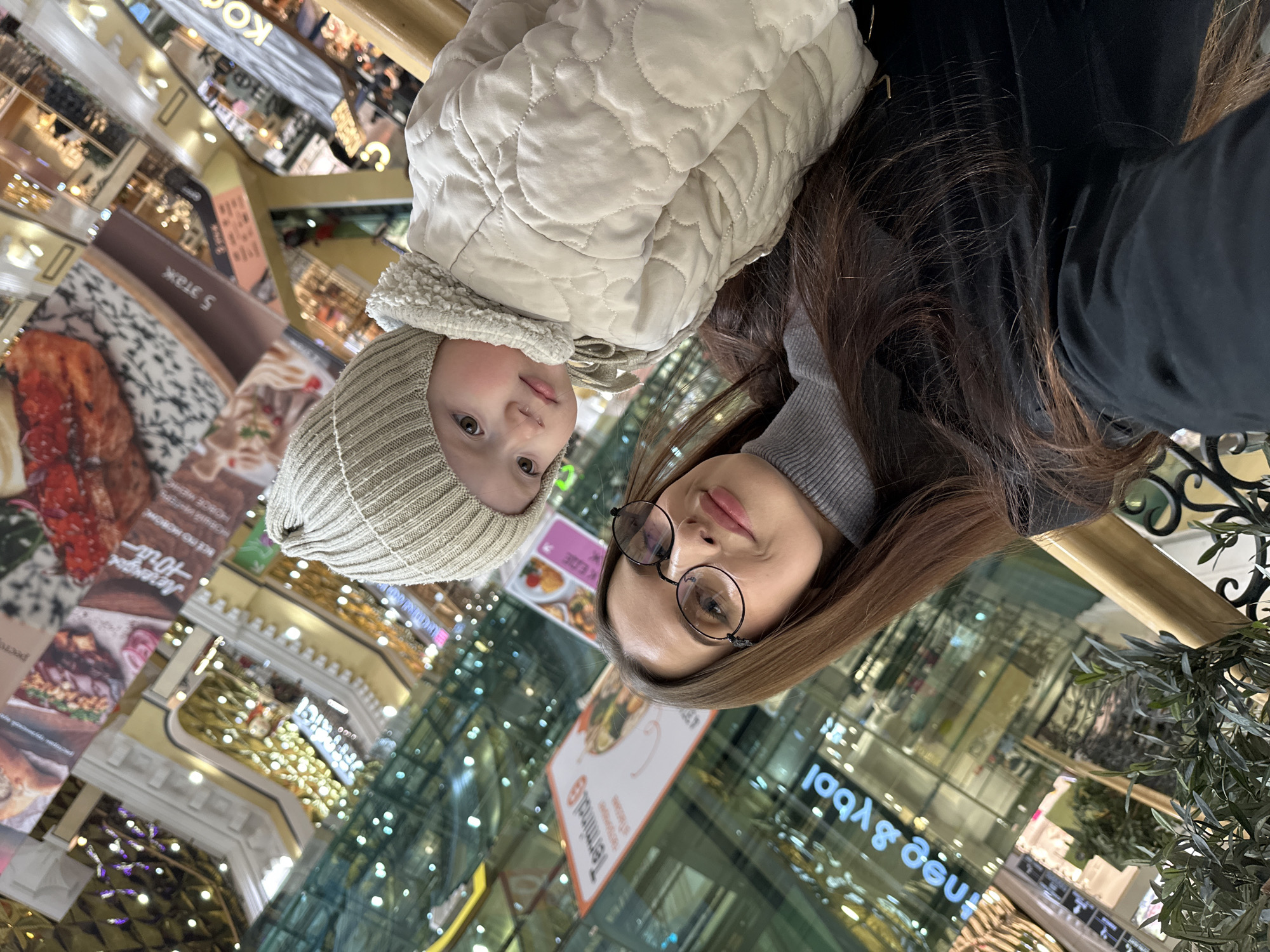 This media file is a rotated, selfie-style photograph, turned approximately 90 degrees counter-clockwise, capturing a woman and a child in a multi-story indoor commercial setting, likely a shopping mall, in Yekaterinburg, Russia.
When viewed upright, the image shows a woman on the right and a child on the left, both looking towards the camera. The woman has long, straight brown hair, wears a grey turtleneck sweater, and round, dark-rimmed glasses. She has a neutral to slightly smiling expression. The child, who appears to be a toddler, wears a light-beige ribbed beanie hat that covers the ears and a puffy, quilted light-beige jacket with a textured, possibly sherpa-like, collar. The child also looks directly at the camera.
The background depicts the expansive, brightly lit interior of a building, characterized by multiple floors, glass railings, and reflective surfaces that create a sense of depth and activity. Numerous artificial lights contribute to a bright, perhaps festive, ambiance. Green foliage from a plant is visible in the lower right portion of the frame.
Visible text in the background includes "TerminalG" and what appears to be "nee & ybol" (possibly a reflected "loop & een" or similar brand name). Other legible Russian text elements include "КЕДЕ," "0000," "Легенды" (Legends), "BCE PO MOBOME" (all new), "ресто" (likely part of "ресторан" - restaurant), and "5 этаж" (5th floor), confirming the multi-level commercial nature of the location. The image conveys a casual outing in a bustling indoor environment.