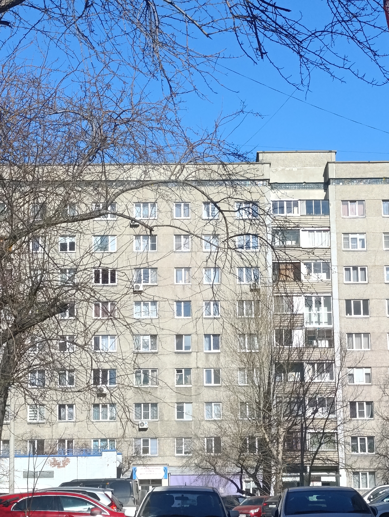 The image shows a residential building in Krasnoyarsk, Russia, likely a panel apartment building common in Soviet-era construction. The building is multi-story and has a gray exterior. Numerous windows are visible across the facade. Bare tree branches are in the foreground, partially obscuring the view of the building and reaching into the clear blue sky. A row of cars are parked in front of the building, indicating a street or parking area. The overall atmosphere suggests a typical urban landscape in the region during a time of year when trees are without leaves.