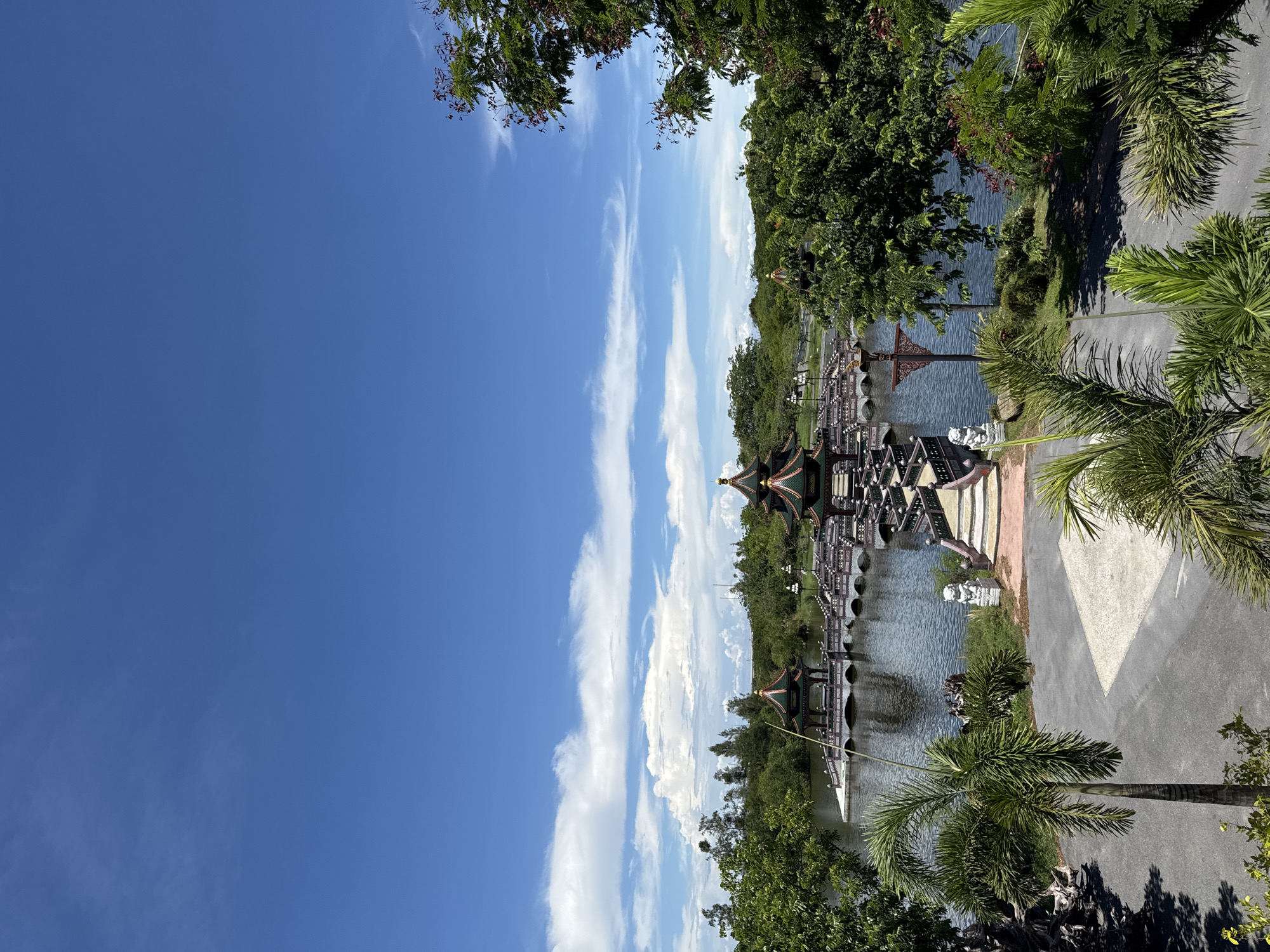 The image depicts a scenic view in Bang Pu Subdistrict Municipality, Thailand. The right side features lush vegetation with various trees, including palm trees, bordering a paved pathway. To the left of the pathway is a body of water, on which a series of intricately designed covered bridges are constructed, connecting several ornate structures. The bridges and buildings exhibit a distinct architectural style, possibly of Chinese or Thai influence, with detailed roofs. The sky, dominating the left portion of the image, is a clear blue with wispy white clouds. No people are visible in the frame.