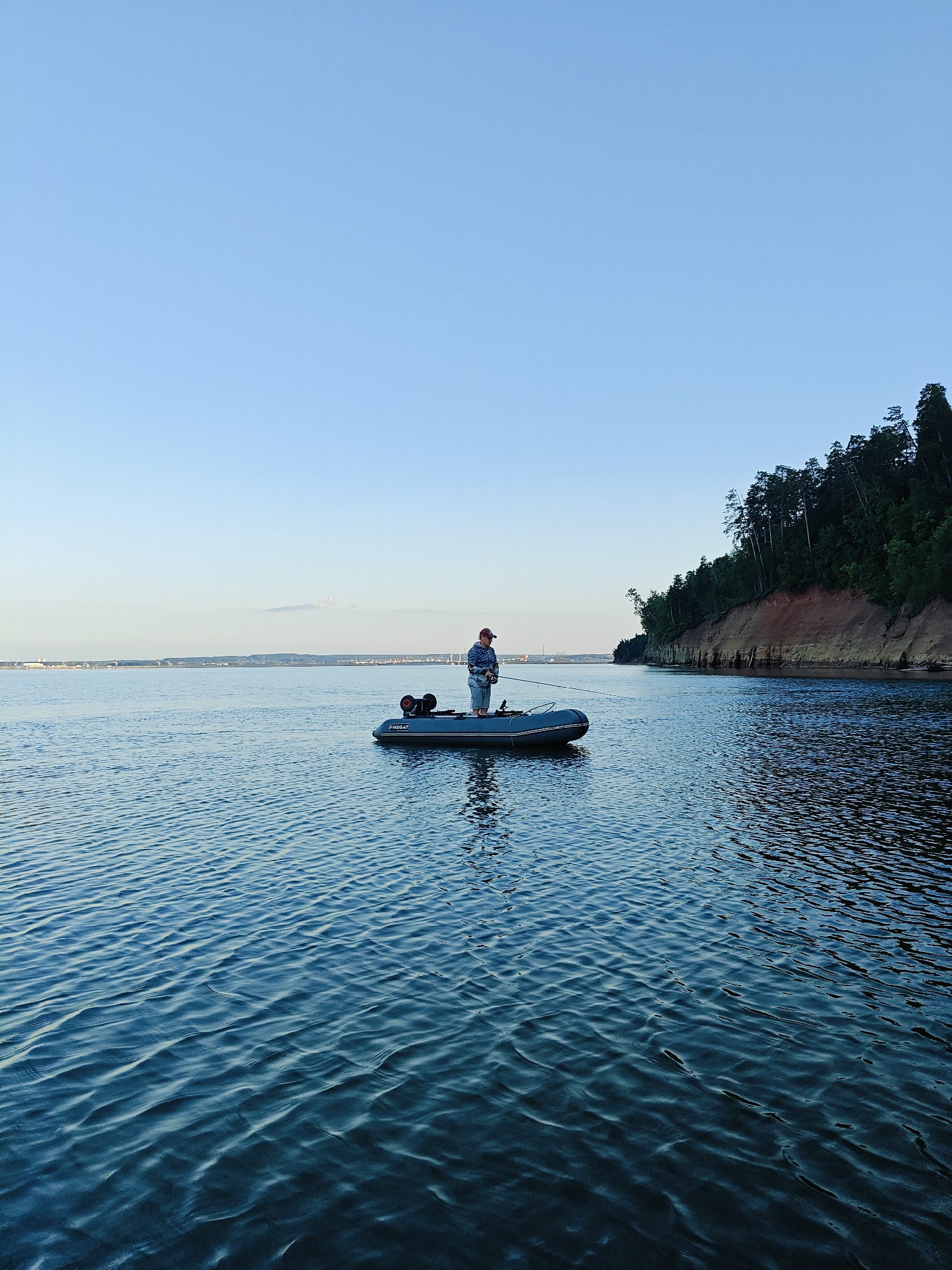 A lone person is fishing from an inflatable boat on a body of water. The person, wearing a red cap and a blue and white patterned shirt, stands in the boat holding a fishing rod. The boat is grey with a black outboard motor at the stern and has the text "FISH CAT" visible on its side.
The scene is set during what appears to be late afternoon or early evening, judging by the soft, warm light and clear blue sky with a faint haze on the horizon. The water is calm with gentle ripples. To the right, a steep, tree-covered bank with exposed reddish-brown soil rises from the water's edge, creating a natural coastline. In the distance, across the water, a hazy shoreline with what appear to be structures or a city skyline is visible under the clear sky.
The overall mood is peaceful and serene, conveying a sense of quiet activity in a natural setting. The location cues, such as the forested bank and the wide expanse of water, are consistent with a river or large lake. The weather appears to be pleasant and calm.