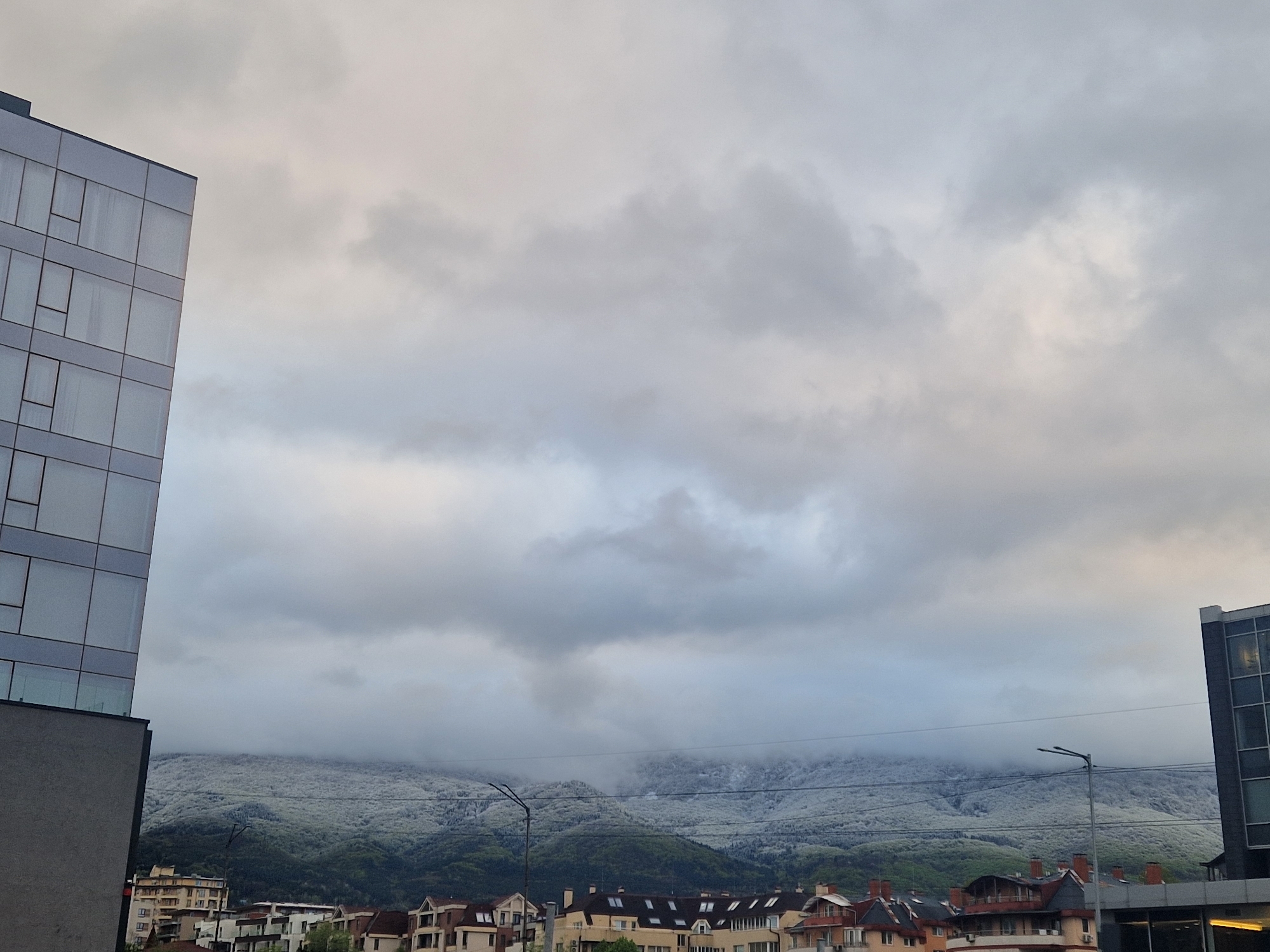 The photograph depicts a city scene in Unknown, Unknown, under a cloudy sky. The foreground features a modern building with reflective glass windows on the left and another building with glass on the right. In the middle ground, there is a row of buildings, including houses with pitched roofs. The background contains a mountain range partially covered in snow or frost, obscured by low-hanging clouds. Two streetlights are visible in the lower center of the frame.