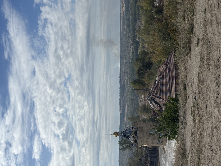 This image captures a panoramic view from an elevated position in Izium, Ukraine. The foreground consists of dry, uneven terrain with sparse light-brown and green vegetation. In the midground, several structures are visible: a prominent, ornate light-colored brick bell tower or chapel, topped with a partially damaged golden dome and cross, revealing a blue-tiled surface beneath. To the right of this structure, a building with a reddish-brown tiled roof shows significant damage, with numerous tiles missing and portions of the roof structure exposed. To the left of the bell tower, a smaller, heavily damaged wooden structure with an exposed, dilapidated roof frame is present. Green and yellowish-brown trees and bushes are interspersed around these buildings and extend into the background. The background features a vast expanse of forested hills and valleys stretching to the horizon. In the distant background, a distinct column of light gray smoke rises vertically before dispersing horizontally. The sky above is partially overcast with numerous white and gray clouds. No individuals are present or discernable in the scene.