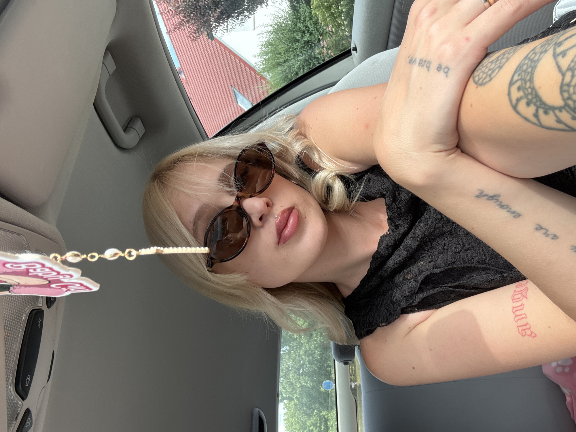 This is a selfie taken from inside a car. The subject is a young woman with blonde hair, wearing sunglasses and a black lace top. She has several tattoos visible on her arms. One tattoo on her left forearm reads "Angel" in red script. On her right arm, there are two tattoos: "be brave" on her upper arm and "are enough" on her forearm. She has a lip piercing.
The setting appears to be daytime, with natural light coming in from the car's windows, suggesting it's not nighttime. Outside the car, trees and a house with a red tiled roof are visible, indicating an outdoor location, possibly a residential area.
A car accessory, a string of beads with a pink cartoon character charm, hangs from the interior of the car, near the rearview mirror. The woman is leaning back, with her arms crossed, and her pose suggests a relaxed or casual mood. The overall image is somewhat tilted, adding a dynamic element to the composition.