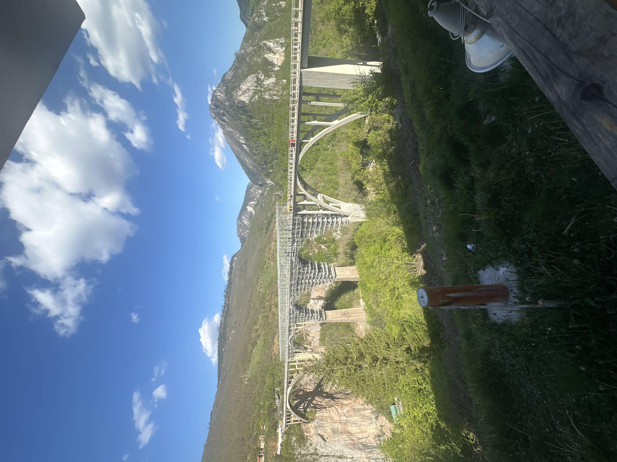 The image captures a view of the Đurđevića Tara Bridge in Montenegro. The bridge spans a deep canyon with the Tara River flowing at the bottom. The mountainous terrain is visible in the background, with a clear blue sky dotted with white clouds overhead. The bridge is a concrete arch structure with multiple spans. Vegetation covers the canyon walls and surrounding landscape. A lamp and a wooden post are visible in the foreground, suggesting the viewpoint is from a structure adjacent to the bridge. The scene appears to be taken during daylight hours.
