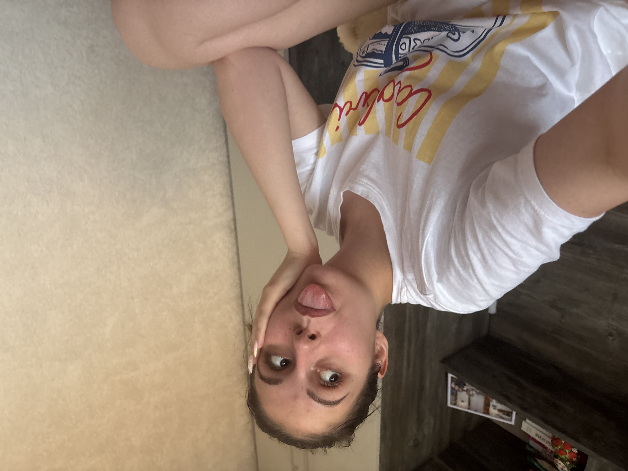 The image presents a fair-skinned young woman wearing a white T-shirt with a print and the word "California" on it. She is sticking her tongue out, with her head tilted sideways and one hand touching her face. The background includes a light-colored carpet and a dark wooden shelf with objects on it. The image does not provide evidence about the location in the city of Bulgarovo, Bulgaria.
