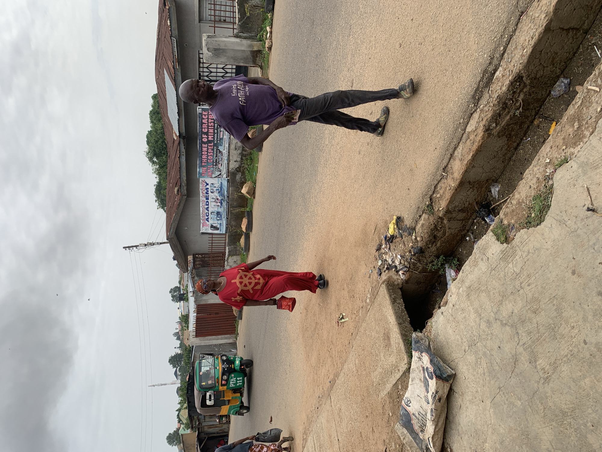 The image shows a street scene in Barakin Akawo, Nigeria. Two men are walking along the side of the road, next to a drainage ditch. One man is wearing a purple t-shirt and dark pants, and the other is wearing a red outfit. A green and yellow auto rickshaw (tuk-tuk) is parked nearby. A building with religious signage is visible in the background. The location appears to be a residential area. The sky is overcast. The street and drainage ditch are littered with trash.