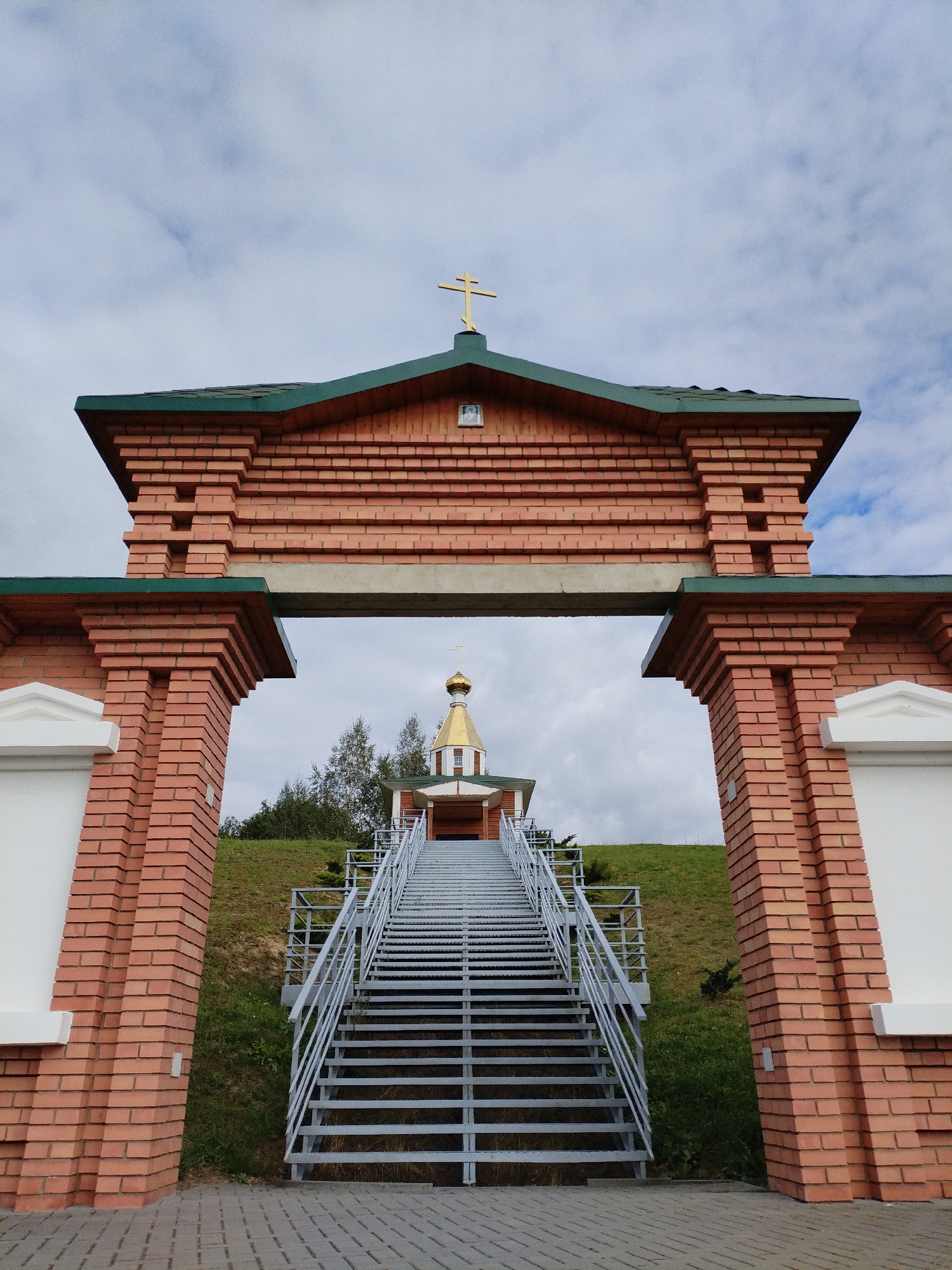 The image shows an outdoor view of a brick archway leading to a set of stairs and a small domed building with a cross. The archway is made of red brick with decorative brickwork and a green roof. The stairs are made of metal with railings. The building appears to be a chapel or small church, also with a golden dome and a cross on top. The background shows a grassy hill with some trees and a cloudy sky. The scene is set in the city of Bialynichy, Belarus. There are no people visible in the image.