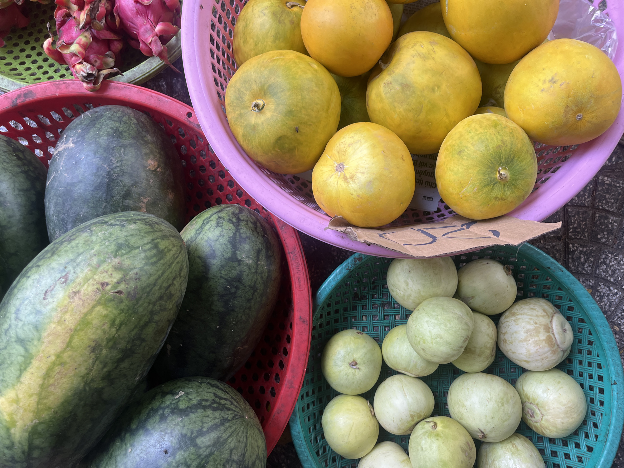 An overhead shot captures a vibrant display of fruits at a street market in Đà Nẵng, Vietnam. In the foreground, a bright pink basket overflows with ripe, yellow melons, their smooth skins catching the light. To the left, a green basket holds a collection of pale green, round fruits, possibly guavas, some showing slight browning. Behind these, a red basket is filled with large, dark green watermelons, their characteristic stripes visible. A corner of another basket on the far right reveals the vibrant pink and green skin of dragon fruit. The fruits are arranged in separate baskets, suggesting they are for sale. The ground beneath the baskets appears to be concrete or pavement, typical of an outdoor market setting. The scene is rich in color and texture, highlighting the abundance of fresh produce available in the bustling city.