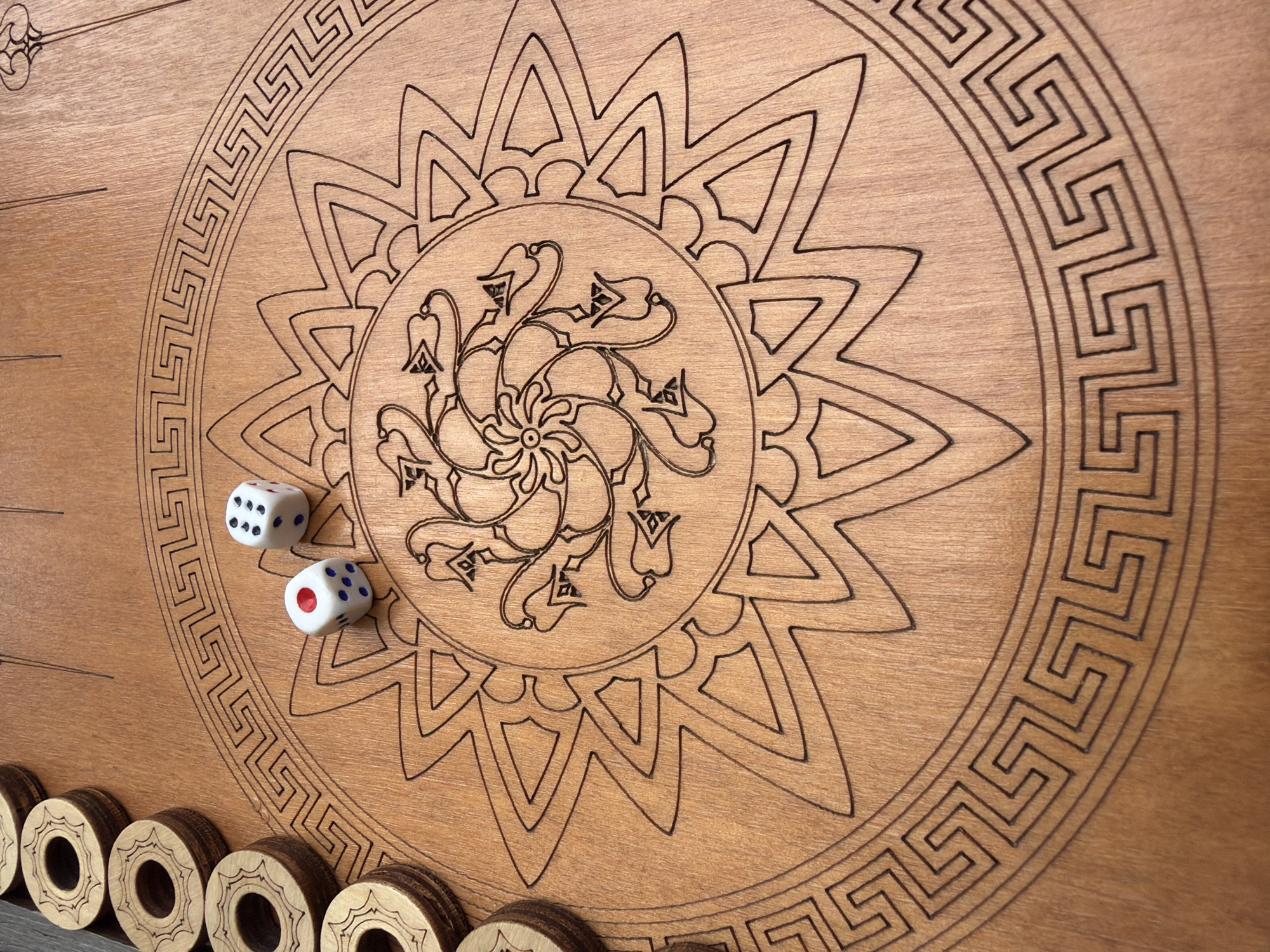 The image shows a wooden backgammon board with dice and checkers. The board is light brown and features a detailed, circular design in the center. The design includes a floral pattern surrounded by a starburst shape and a Greek key border. Two dice are positioned on the board, one showing 5 and 1 (6), and the other showing 1 and 5 (6). A row of wooden checkers with darker brown edges is located along the bottom edge of the board. The scene is tightly framed and lacks information to determine the location in Budva, Montenegro.