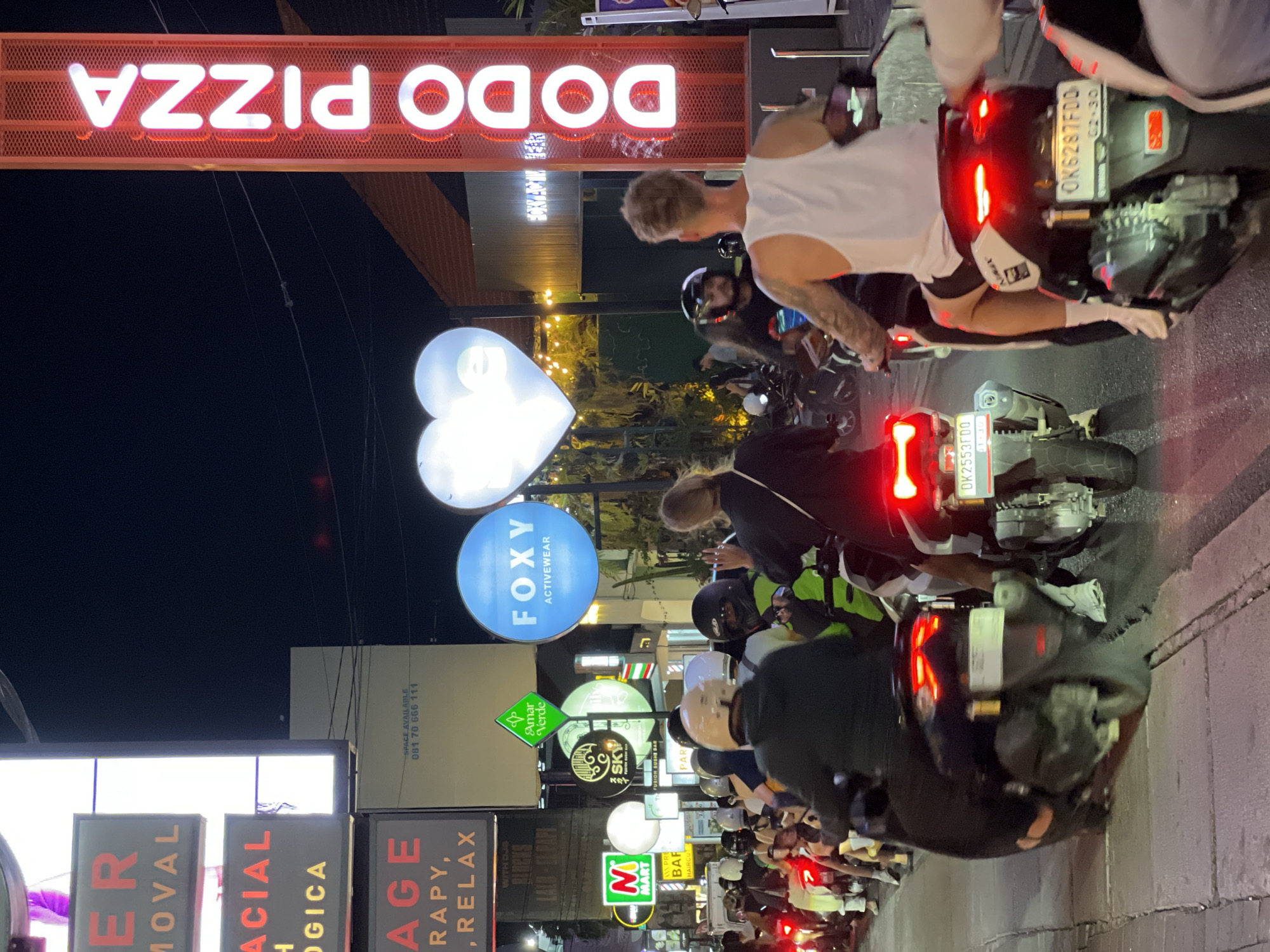 The image captures a busy street scene at night in North Kuta, Indonesia. A long line of scooters and motorcycles is seen on the right side of the road, with people riding them. Various businesses are lined along the street, indicated by numerous neon signs and advertisements. Notable signs include "DODO PIZZA", "FOXY ACTIVEWEAR", and several promoting massage and therapy services. A heart-shaped light fixture is also visible. The street is brightly lit by the shop signs and vehicle lights.