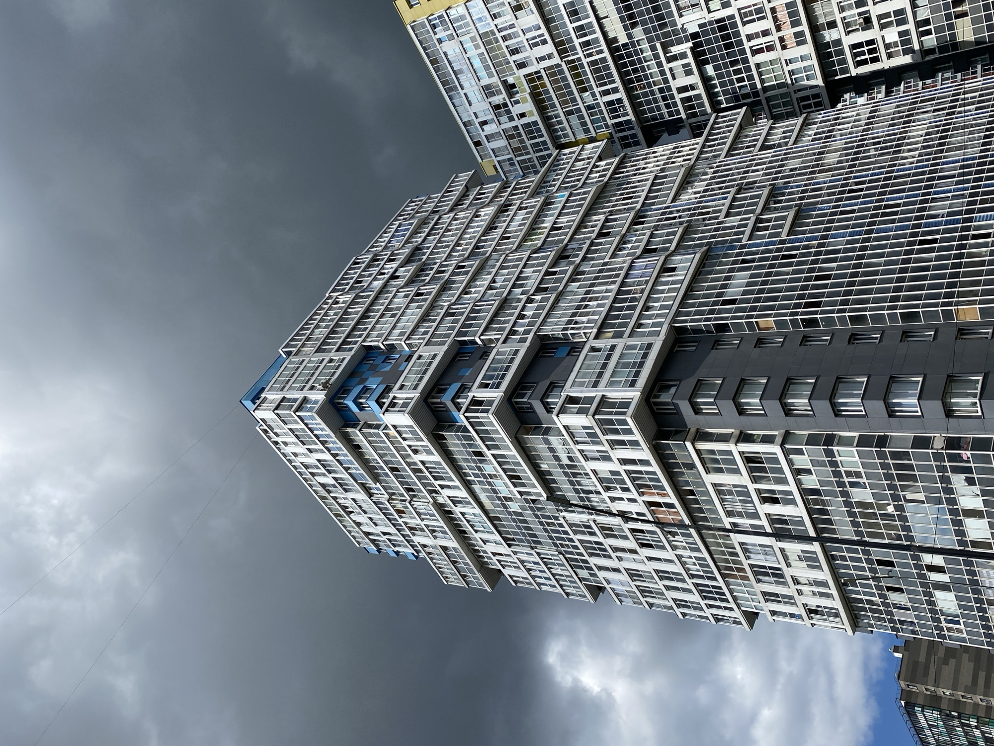Two tall, modern residential buildings are viewed from a low angle against a dark, cloudy sky in Kudrovo, Russia. The building on the left is primarily white with gray accents and numerous windows. It features a staggered facade with protruding sections. Blue architectural elements are visible on the upper floors. The building on the right, partially visible, has a yellow upper section and a facade predominantly of white and gray panels with many windows. The sky is overcast with dark, heavy clouds, suggesting impending rain or a stormy day. No people or specific actions are discernible in the image. The environment is urban, characterized by the presence of large apartment complexes.