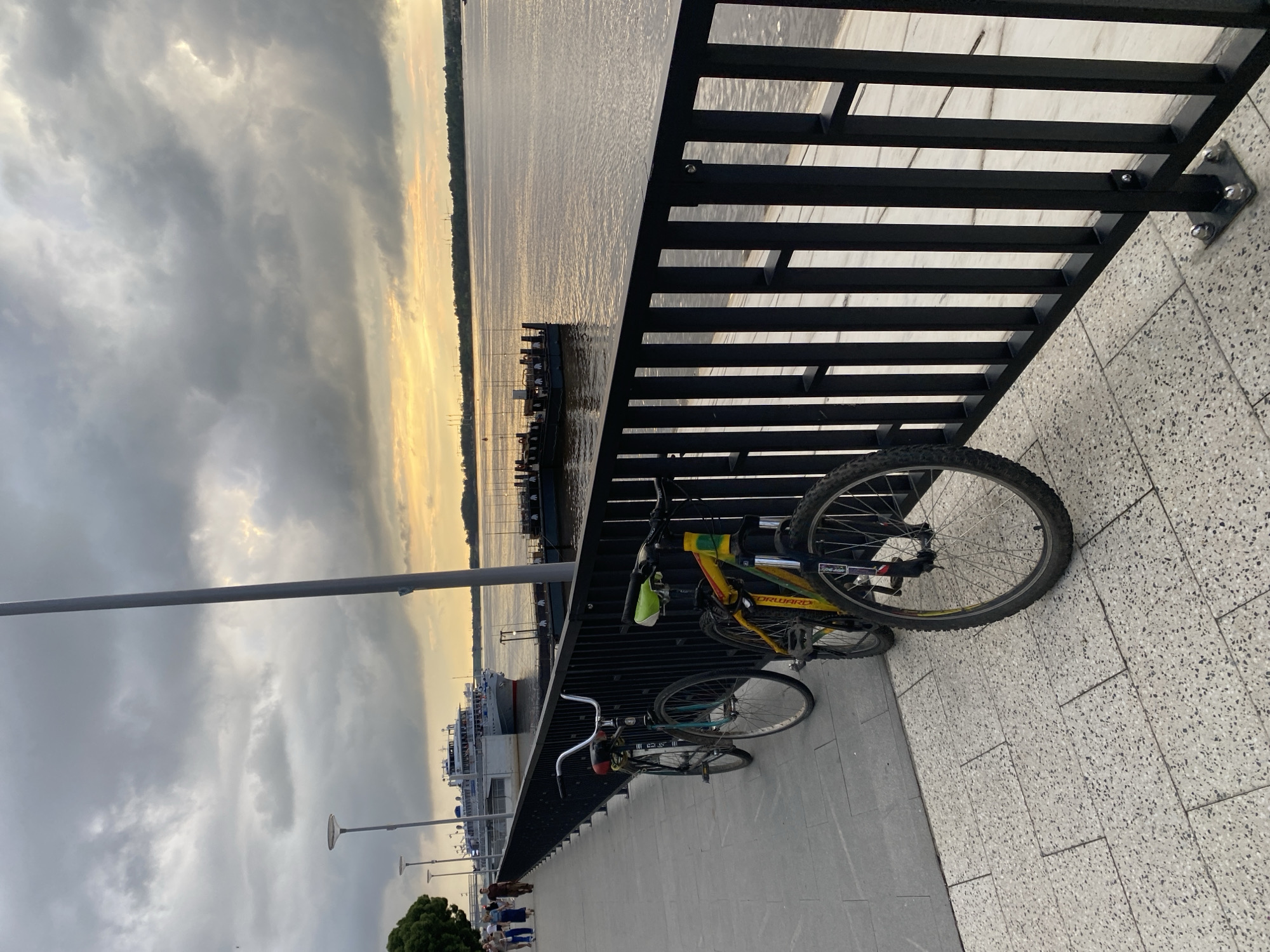 This image depicts a waterfront promenade in Perm, Russia, likely during overcast weather with a sunset occurring over the Kama River. Two bicycles are parked on the paved walkway, leaning against a black metal railing. Beyond the railing, the river stretches towards the horizon, with a large passenger boat docked and several smaller structures visible on the water. The promenade itself is lined with lampposts, and a few individuals are walking in the distance. The ground is paved with textured tiles.