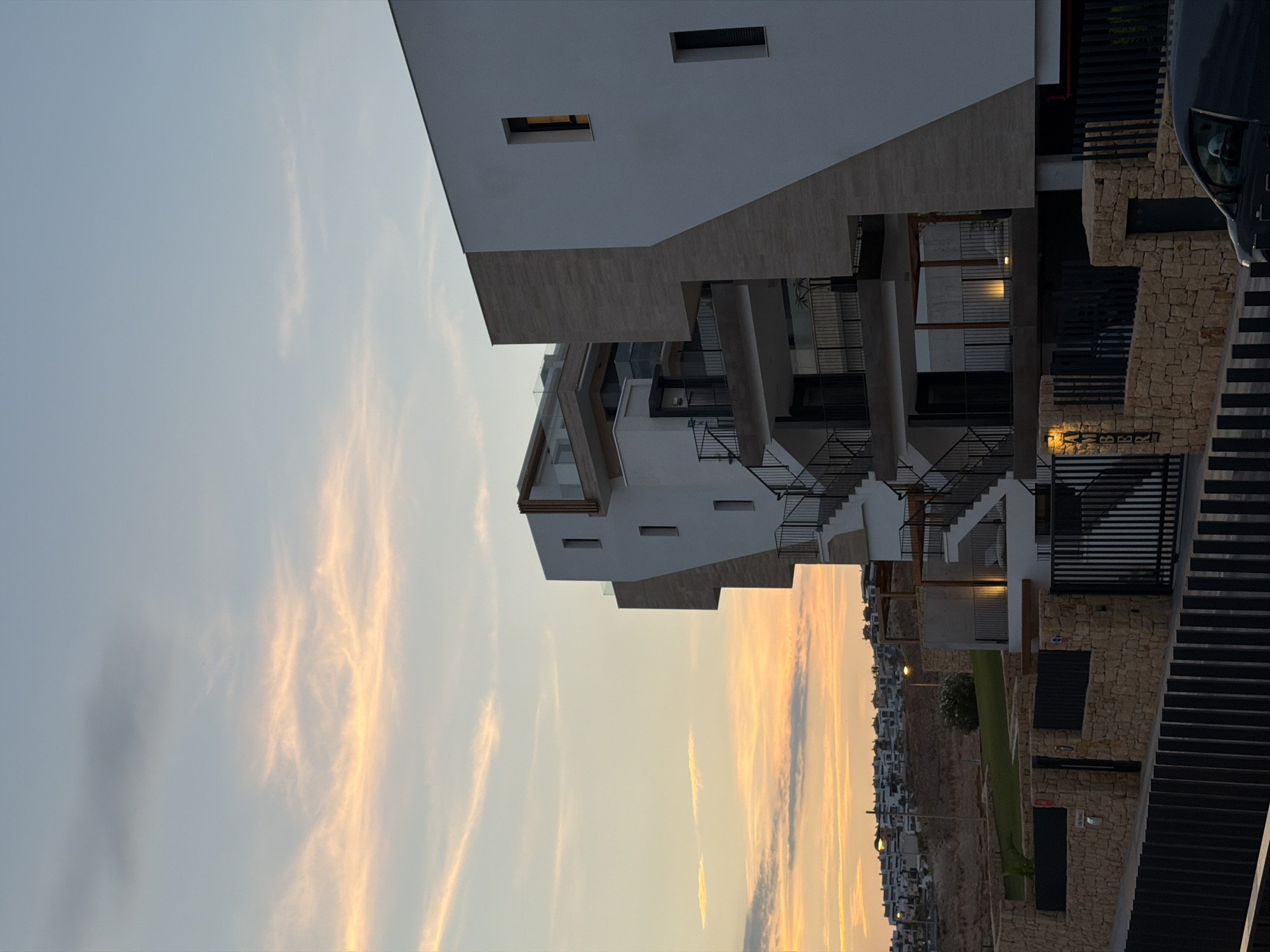 This is a medium-angle, vertical shot of a modern multi-story building in Finestrat, Spain, during a sunset. The sky is a gradient of pale blue to soft orange and pink, with wispy clouds. The building itself is white with geometric angles and accents of light-colored stone. It features multiple balconies with black railings and external staircases. One of the walls has a large, asymmetrical diagonal line of stone.
In the background, a hillside is dotted with numerous white houses, suggesting a developed urban area. The foreground shows a low stone wall and a black metal fence, with a street and the front of the building's ground floor visible. A dark-colored vehicle is parked on the right side of the frame, partially obscured by the fence. The name "AMBER" is visible on a lit sign attached to a stone pillar near the fence. No people are visible in the image.