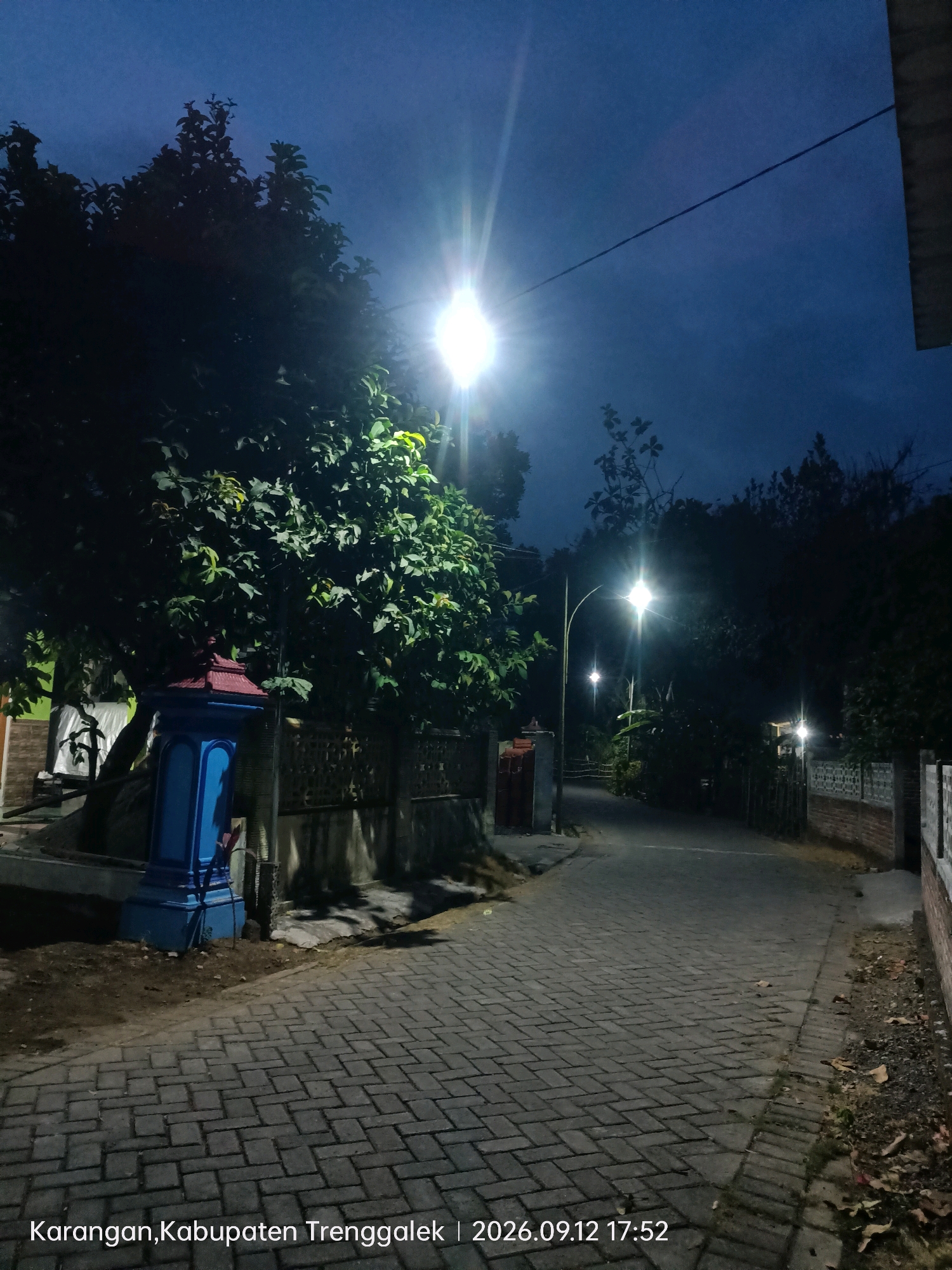 The image depicts a paved pathway or road at dusk or night, illuminated by streetlights. On the left side of the pathway, there is a large tree with dense foliage and a blue and red structure, possibly a post or small building. The pathway is made of interlocking bricks and extends into the distance, where more trees and streetlights are visible. The sky is a deep blue, indicating low light conditions. The foreground shows the brickwork of the path, with some scattered leaves. A timestamp and location, "2026.09.12 17:52 Karangan, Kabupaten Trenggalek", are overlaid on the bottom of the image.