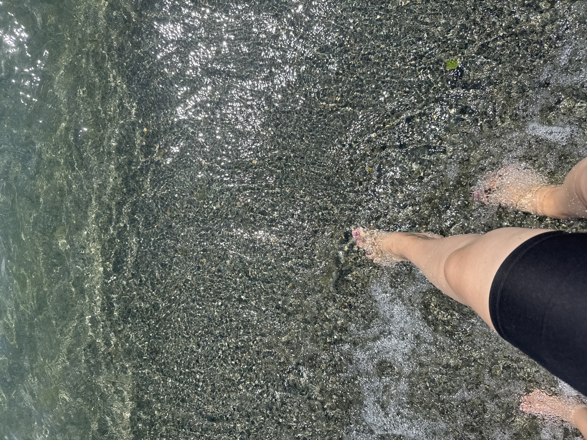 A person's legs and feet are visible, submerged in shallow, clear water. The water's surface is disturbed by ripples and reflections of sunlight. The bottom of the water body is covered in small, dark pebbles. The person is wearing black shorts that extend to mid-thigh. The scene appears to be a natural water environment, possibly a lake or shallow sea, given the pebbly shore and clear water. The location is identified as Tsamakaberd, Armenia, suggesting a natural area within or near the city.