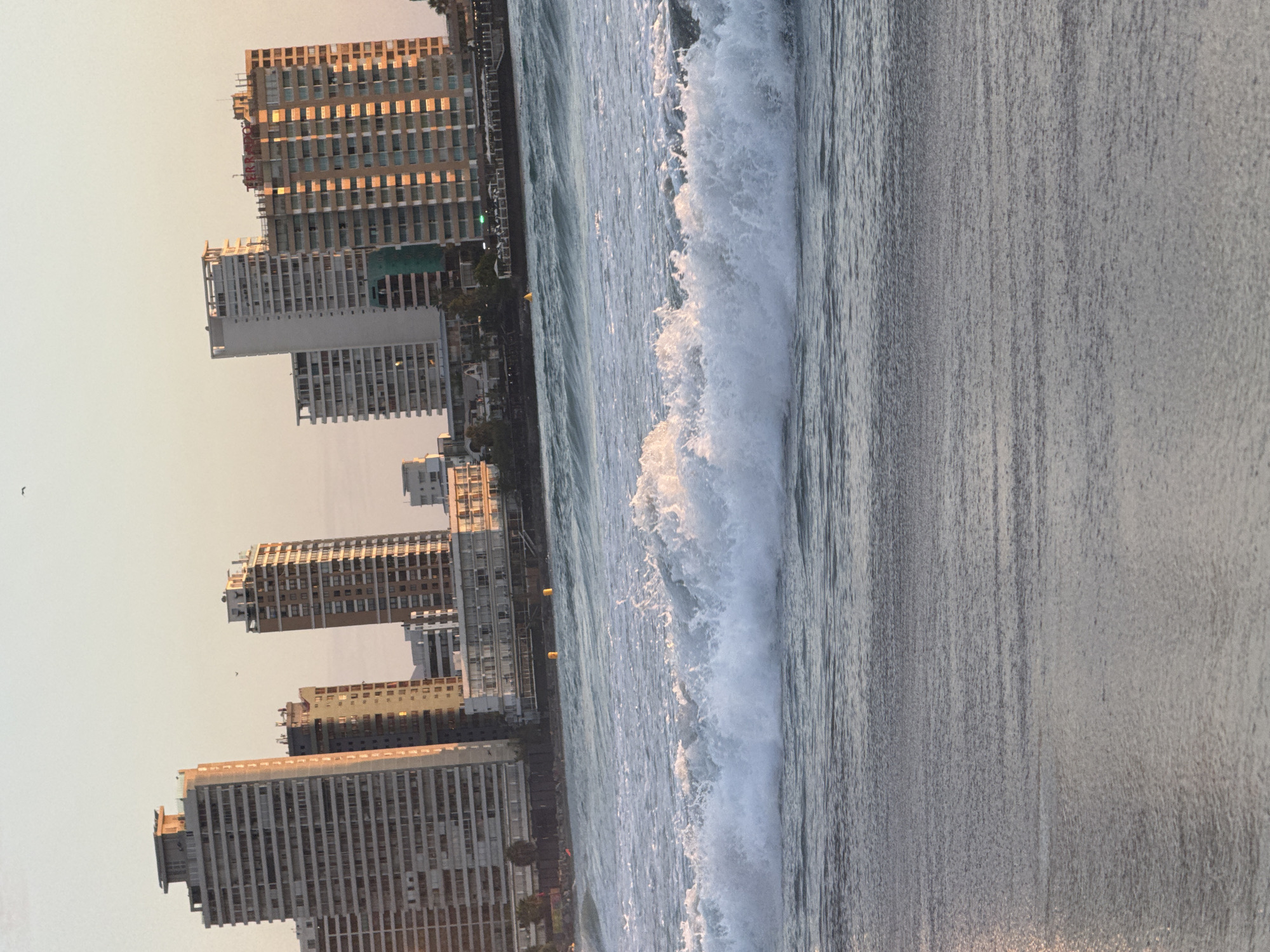 The image depicts a beach scene in an urban setting, with a row of high-rise buildings visible along the shoreline. Waves are crashing on the beach, creating a line of white foam. The water appears calm beyond the breaking waves. The buildings are a mix of architectural styles, with some appearing older and others more modern. The sky is a pale color, suggesting either early morning or late afternoon light. The location is Unknown, Unknown.