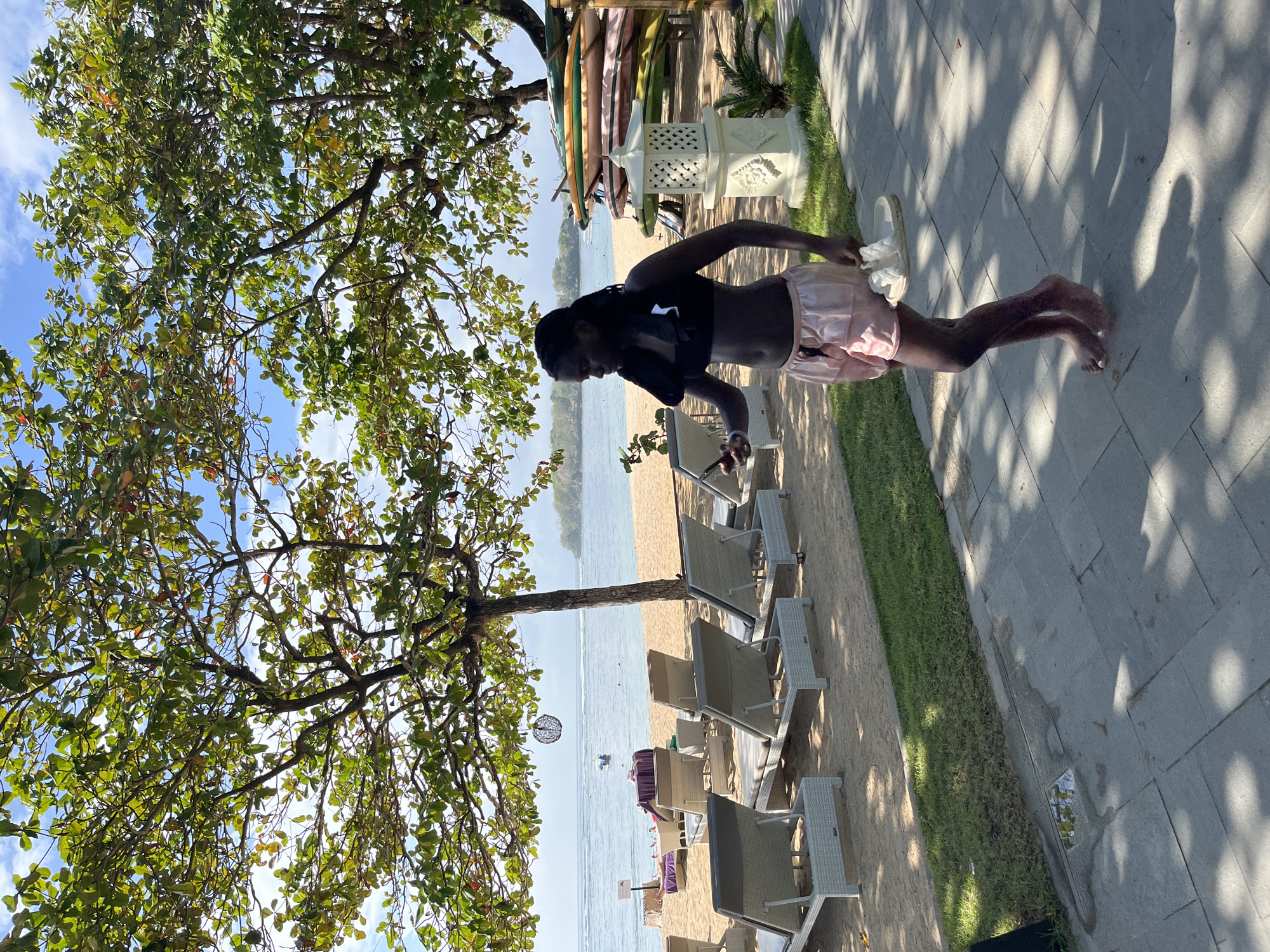 A Black woman wearing a black bikini top and pink silk shorts walks along a paved pathway next to a grassy area. She is holding a white sandal in her right hand and a white object in her left hand. Behind her, a sandy beach is lined with lounge chairs and umbrellas. The ocean is visible in the background with a few boats on the water. Several surfboards are stacked vertically on the beach to the right of the woman. A white decorative lantern sits on a pedestal to her right. The scene appears to be a resort or beach club in Kuta Selatan, Indonesia, characterized by tropical trees and clear blue skies.