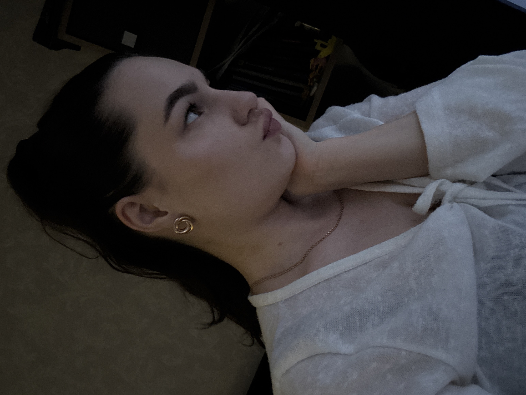 A young woman, with dark hair pulled back and a white top, is pictured in a close-up, profile shot. Her head is tilted upwards and slightly to the left, with her right hand supporting her chin. She is wearing small, circular gold earrings and a delicate gold chain necklace. The background is dark and out of focus, suggesting an indoor setting, possibly at night or in low light conditions. The overall mood appears contemplative or pensive. There is no visible text in the image. The location is Chaikovsky, Russia.