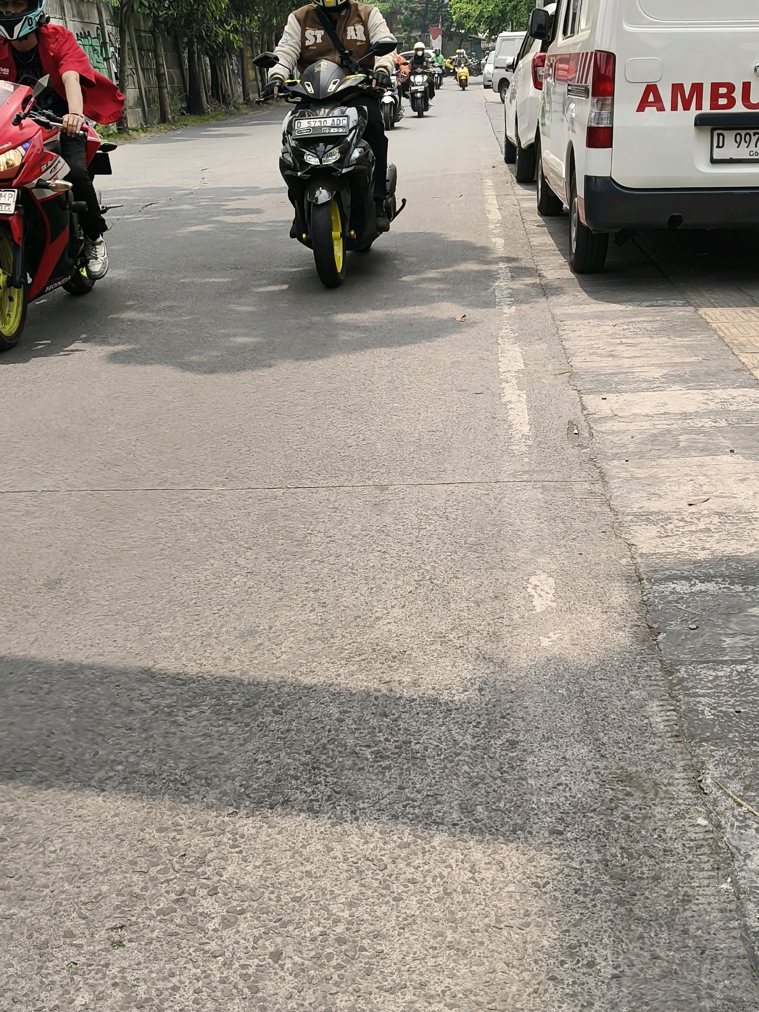 This is a medium-angle, eye-level shot of a street scene in Bandung City, Indonesia. The foreground features the asphalt road with a light gray and dark gray speckled texture, divided by a subtle horizontal line. A large, soft shadow from an unseen object is cast across the left side of the road.
In the middle ground, two motorcycles are prominent. The one on the left is red with yellow accents on its wheels. The rider, wearing a red jacket and black pants, is partially visible from the waist up. The motorcycle on the right is black with yellow accents on its wheels and a license plate reading "D 6730 ADC". The rider is wearing a brown and white jacket and black pants, with a helmet on.
Further in the background, a white ambulance with red lettering that partially reads "AMBU" is parked on the right side of the road. Another white van is positioned in front of it. Several other motorcycles and a yellow scooter are visible further down the street, suggesting moderate traffic. The background includes a concrete wall with graffiti and trees lining the street, indicating an urban or semi-urban environment. The lighting suggests it is daytime, likely sunny due to the distinct shadows.