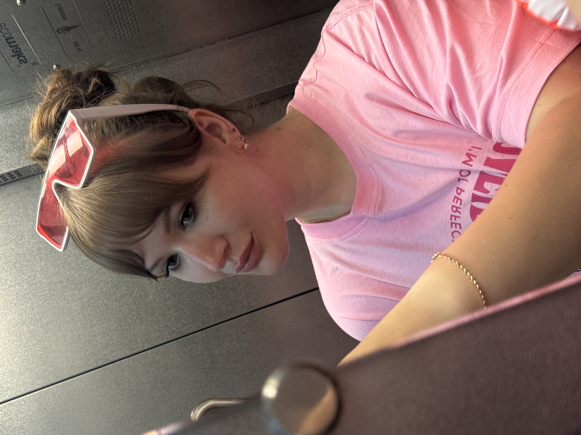 A young woman with auburn hair is looking down and to the left, wearing pink framed sunglasses on her head, a gold necklace, a pink T-shirt with text in reverse, and a gold bracelet on her left arm. She is in an elevator, pressing a button. The elevator has gray metal walls. The location is unknown, and there is no verifiable evidence suggesting it is in Yevpatoriya, Ukraine.