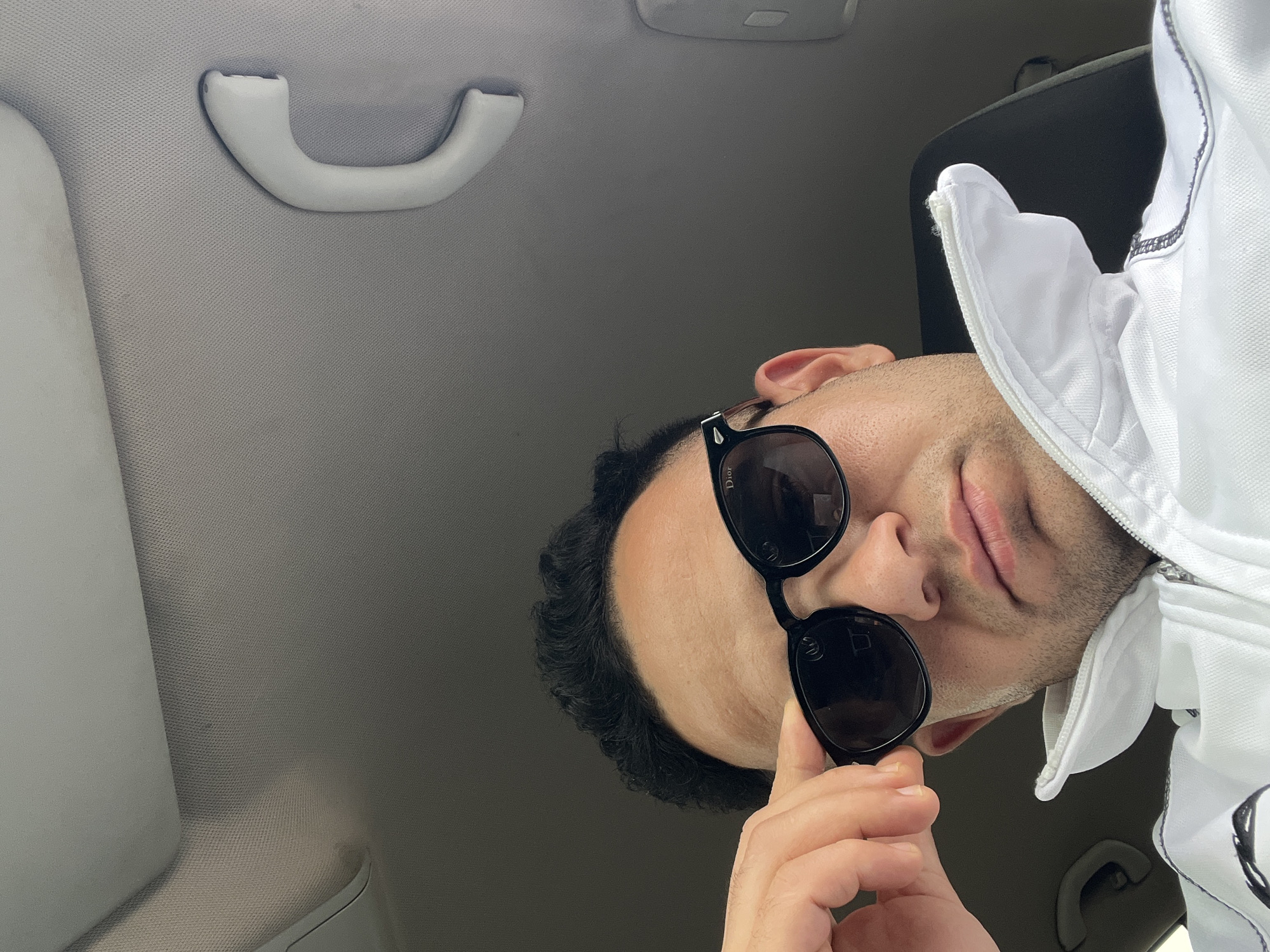 This image offers a unique, low-angle perspective looking upwards at a man seated inside a car, likely in Martil, Morocco. The man's head and upper chest are visible, with his face slightly tilted towards the right. He is wearing dark sunglasses, prominently displaying the "Dior" brand name on the temple, and a white hooded jacket with a zipper and black stitching details on the hood's edge. His dark, wavy hair frames his face, and a light mustache and beard are noticeable. His right hand is raised, with fingers gently resting near the frame of his sunglasses. His expression appears neutral or slightly relaxed, with his lips slightly parted.
The background clearly depicts the interior of a car, dominated by the grey, textured fabric of the headliner. A white overhead grab handle is visible in the upper left corner, along with parts of the car's ceiling structure and what appears to be a sun visor. The lighting suggests it is daytime. The photograph is composed in a way that implies a selfie or a shot taken from a very low vantage point, perhaps from a passenger seat or dashboard level, looking up at the driver or front passenger.