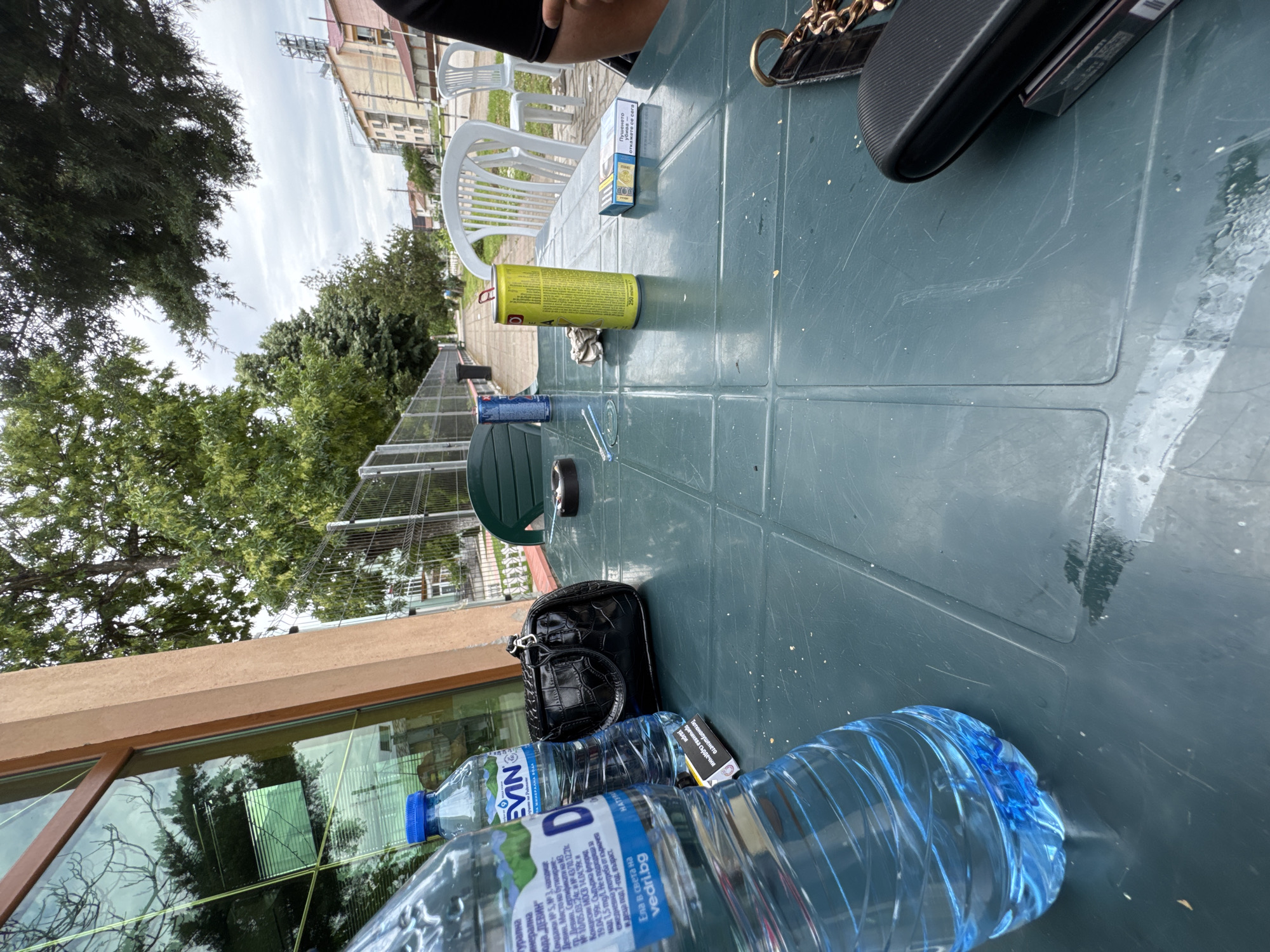 The image shows a green plastic table cluttered with various items. Two large bottles of "Devin" mineral water, one with a blue cap, are prominent in the foreground. A black purse and a package of cigarettes are positioned nearby. A yellow can, possibly an insecticide or cleaning product, lies further up the table, along with a pair of glasses and a roll of black tape. In the background, there are white plastic chairs and glimpses of greenery, including trees and a fence. The location appears to be outdoors, possibly a patio or garden, with a building visible in the distance, suggesting a setting within the city of Drenovets, Bulgaria, based on the water bottle brand.
