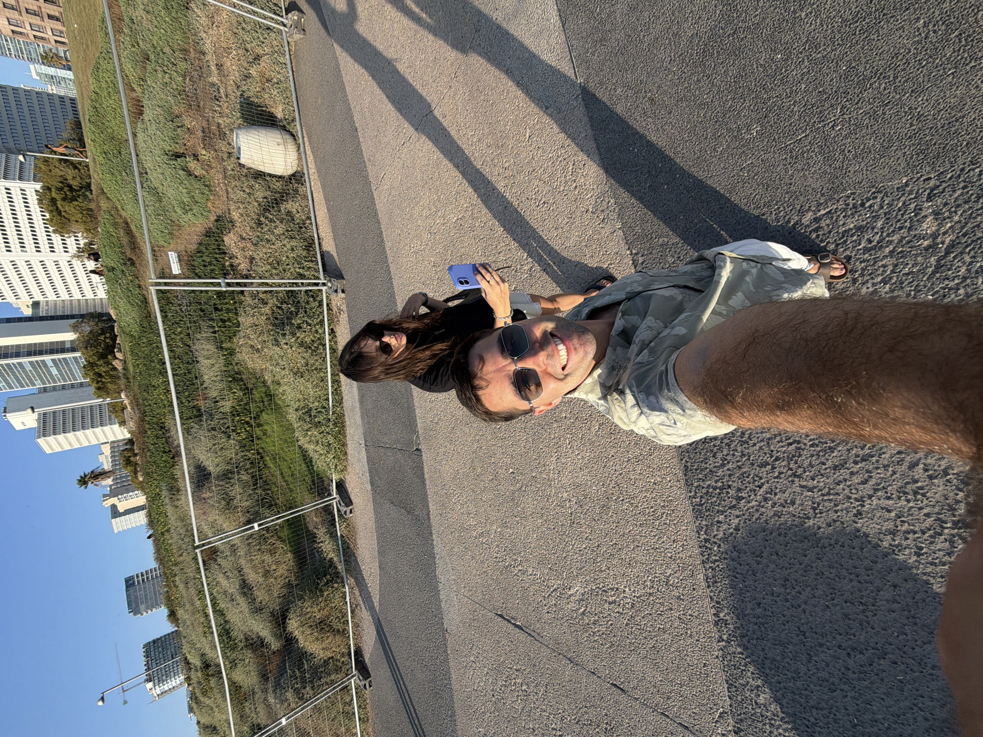In this high-angle, full-body selfie captured outdoors in Tel Aviv, Israel, a man and a woman are standing on an asphalt path with a slightly sloped, vegetated embankment behind them. The embankment is lined with a temporary wire fence and features scattered bushes and grasses. In the background, a cluster of modern, white high-rise buildings dominates the skyline under a clear blue sky. The man, in the foreground, is wearing sunglasses and a light-colored, patterned short-sleeved shirt. He is smiling directly at the camera. The woman, positioned behind him and slightly to his left, is also wearing sunglasses and a black top, holding a purple smartphone in her left hand as if taking a photo or video. Her right hand is visible by her side. Both are dressed casually for warm weather, with the man wearing shorts and sandals, and the woman appearing to wear a skirt or shorts. Long shadows from unseen objects stretch across the asphalt path, indicating it is late afternoon or early morning. The scene appears to be a public space, possibly a park or promenade, adjacent to the urban landscape.