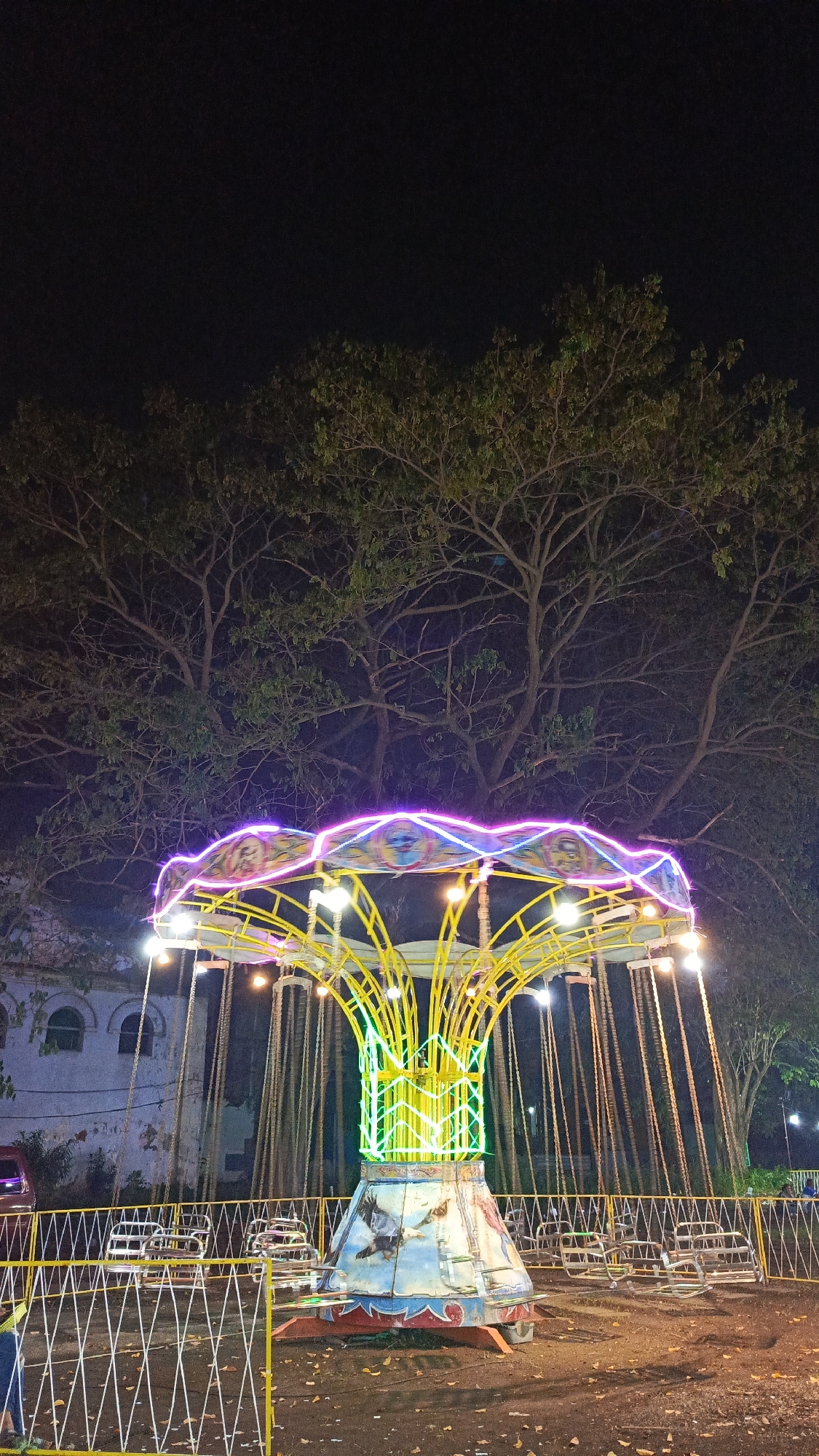 The image shows a swing carousel illuminated with neon lights at night. The ride has a yellow metal frame, a central green neon structure, and swing seats suspended by chains. The carousel's top has alternating yellow, pink, and blue lights, and its base is decorated with a painting of a bird. A white picket fence surrounds the ride, and the ground is covered with fallen leaves and debris. Bare trees with sparse foliage are visible in the background, along with a building with arched windows. A small portion of a red vehicle can be seen on the left edge of the frame, suggesting a street. The location is Tulangan, Indonesia.