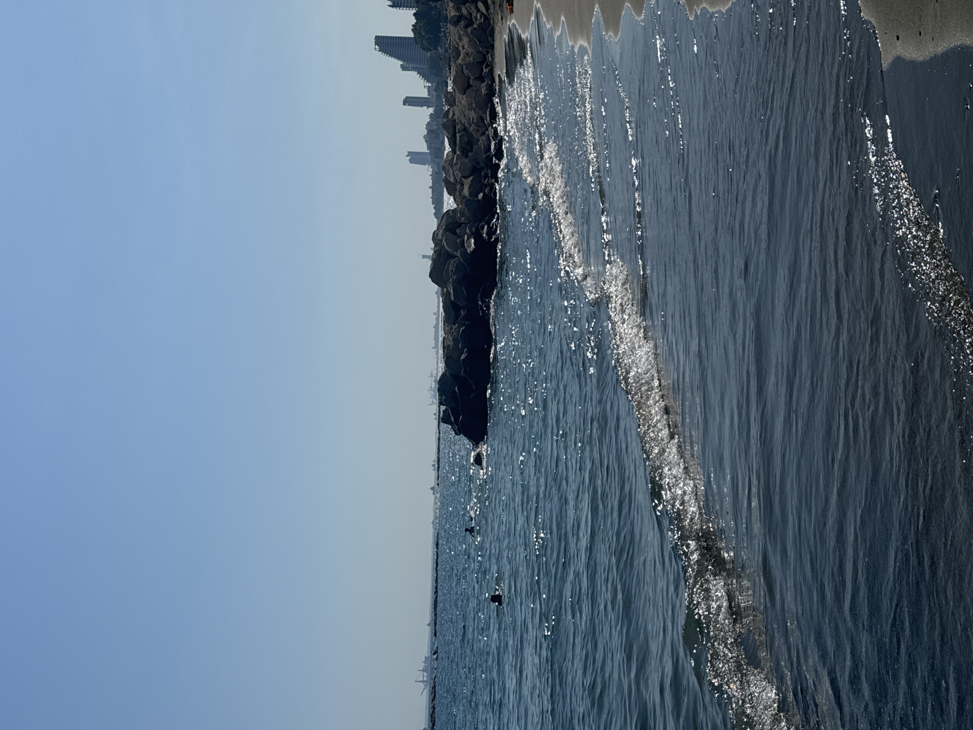 The image depicts a coastal scene in Agios Tychona Community, Cyprus. The foreground features the dark blue sea with small waves gently lapping at a sandy beach. Sunlight reflects off the water's surface, creating shimmering highlights. In the mid-ground, several large, dark rocks form a breakwater, with a few people visible swimming in the water around them. Beyond the rocks, the coastline stretches out, lined with modern, high-rise buildings that form a cityscape against the clear blue sky. In the far distance, several large ships and industrial structures are visible on the horizon, suggesting the presence of a port or harbor. The overall impression is of a sunny day at a developed coastal area.