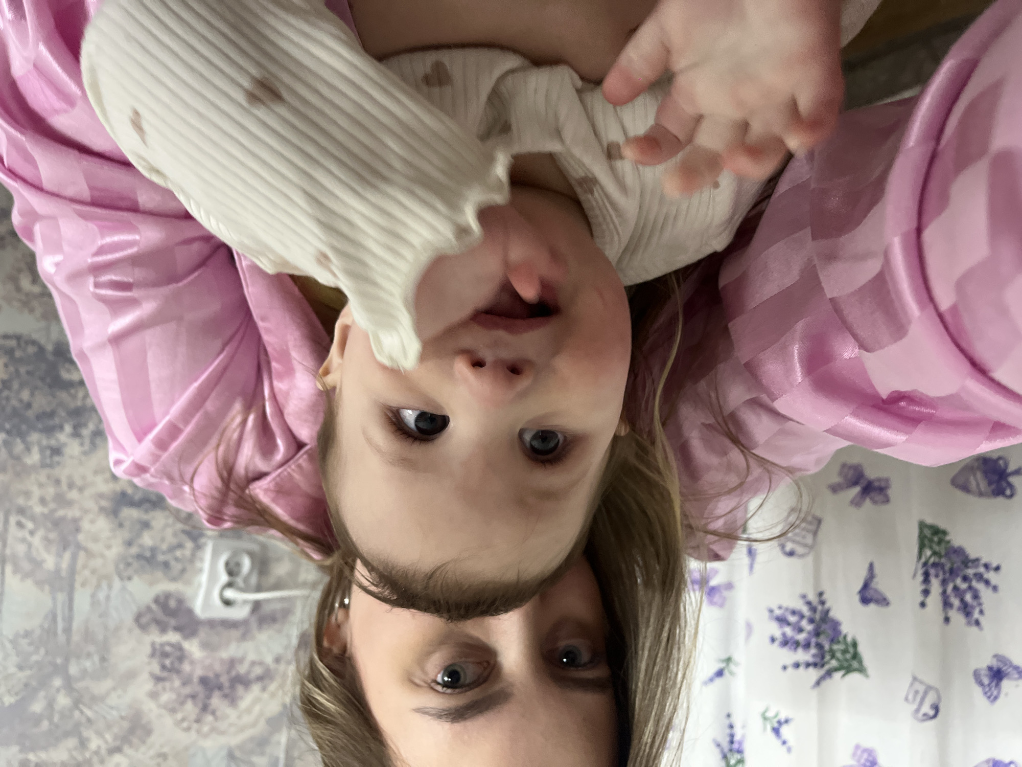 A rotated image shows a young girl and a woman's face. The girl is wearing a white sweater with small heart patterns and has her mouth slightly open. The woman is positioned below her. Both have light skin and similar eye color. The woman's hair is light brown. The background includes a pink striped fabric, possibly a robe, and a wall with a floral pattern. An electrical outlet is visible on the wall. The scene appears to be indoors. There is no visible information to determine the location to be the city of Novopokrovskaya, Russia.