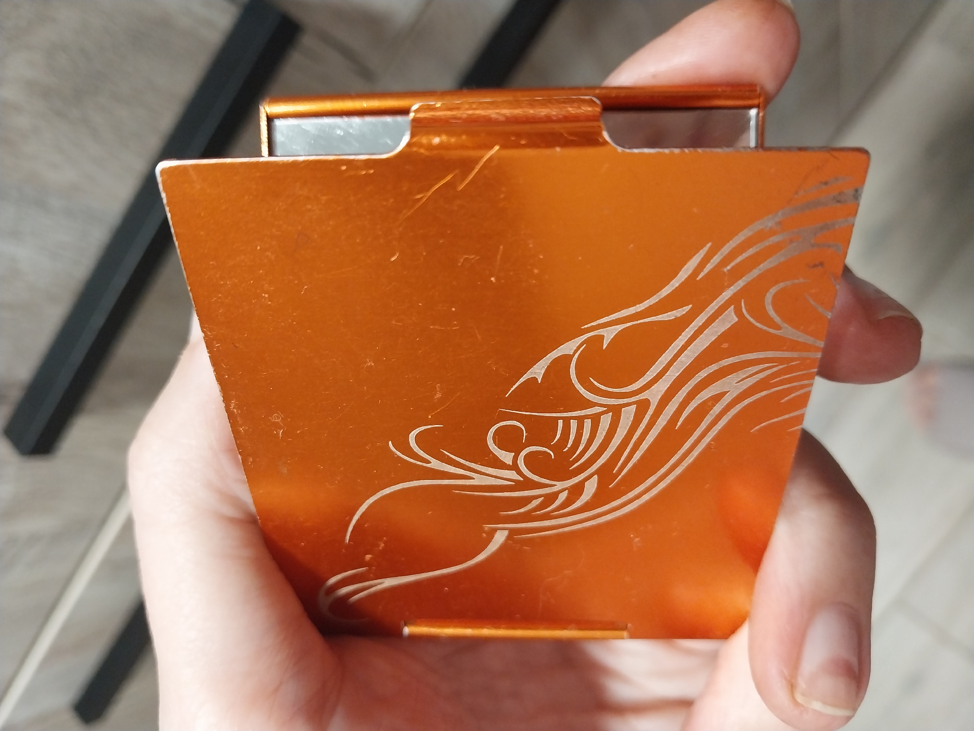 A person's hand is holding an open, orange, metallic cigarette case. The case has a decorative, swirling white pattern etched into its surface, resembling flames or abstract floral designs. The case appears to be made of aluminum. The background is blurred and shows a wooden surface with black horizontal elements, possibly shelves or drawers. The scene is indoors. No specific location in Dnipro, Ukraine can be identified from the provided image.