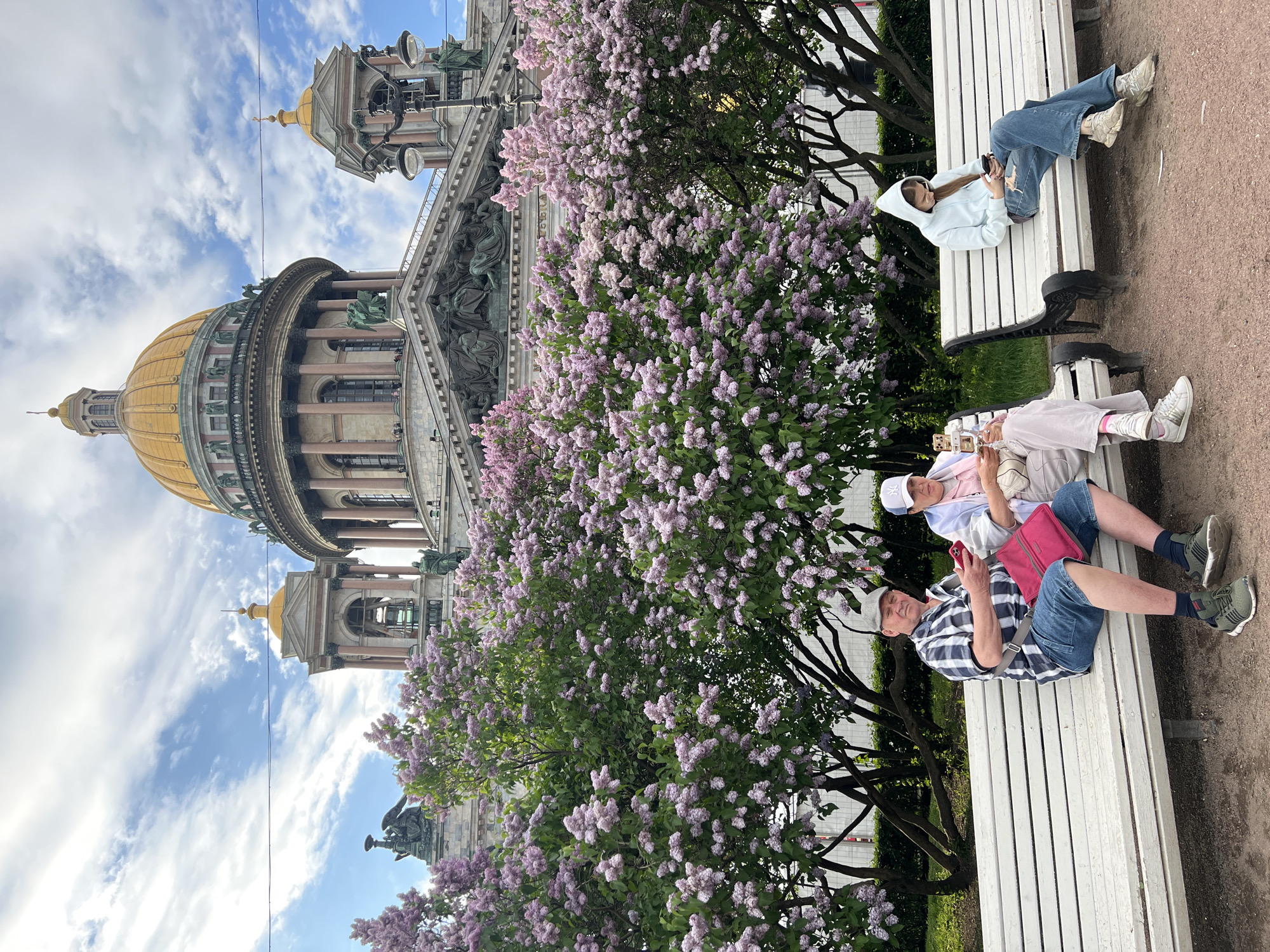 The image depicts a group of people relaxing on white benches in front of a large building with a golden dome, possibly a cathedral or major landmark. Lilac bushes in full bloom are prominent in the foreground, partially obscuring the building. Three people are seated on the bottom bench: a man in a plaid shirt and denim shorts, a person in a pink outfit and cap, and another individual partly obscured. On the upper bench, a person wearing a white hooded sweatshirt and jeans is lying down, looking at their phone. The building behind them appears to be a historical structure with ornate details, statues, and pillars. The sky is visible in the background, with clouds. The location appears to be a park or public space in a city.