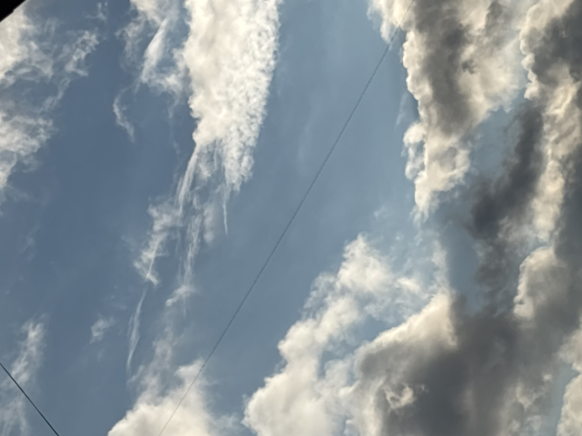 The image shows a sky with clouds. Some clouds are white and wispy, while others are larger and darker gray. A faint contrail runs diagonally across the blue sky. The exact location in Perm, Russia, is not discernible from the image. There are no people, specific objects beyond the clouds and contrail, or any discernible actions or interactions in the image.