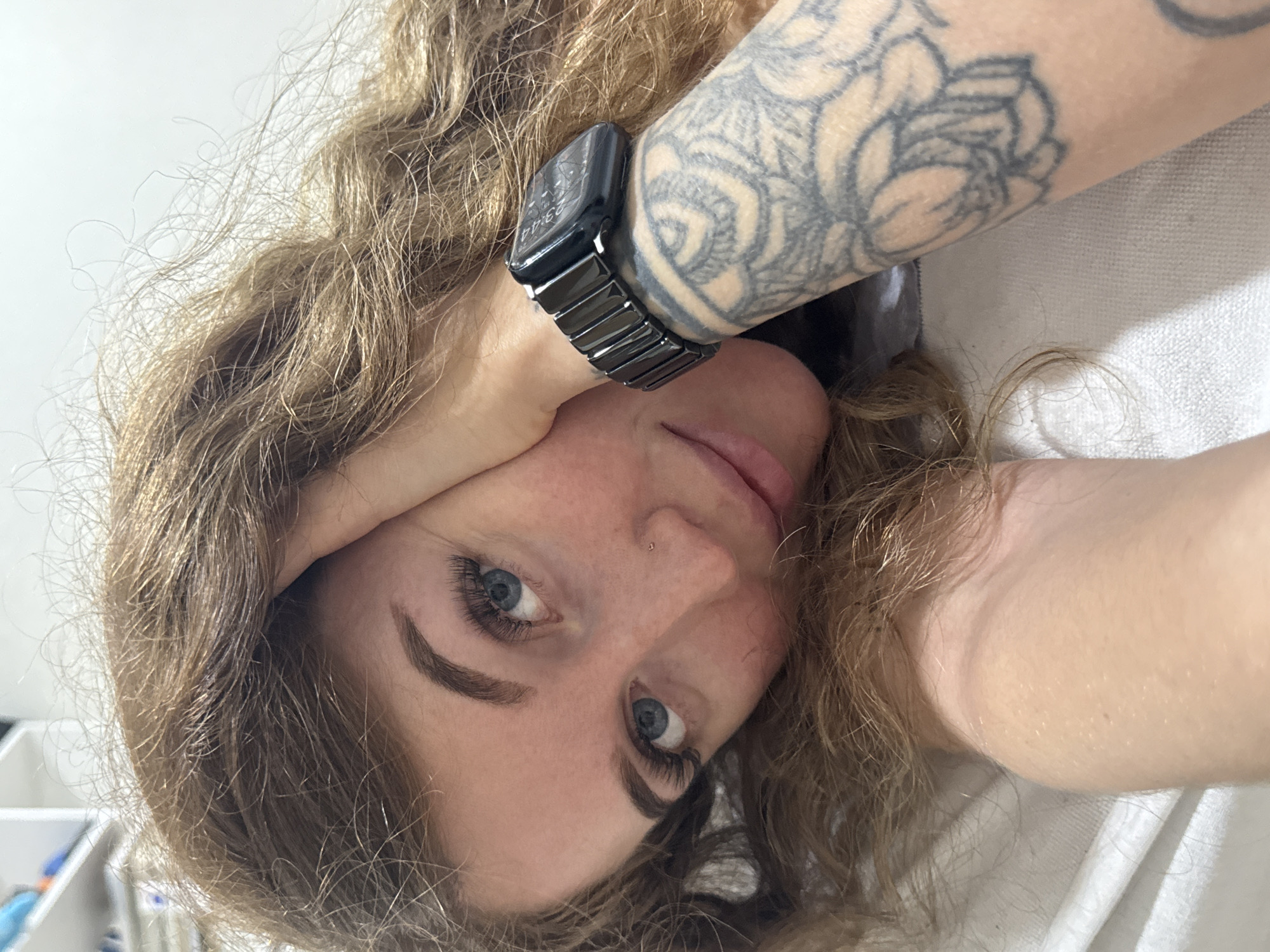 A selfie of a fair-skinned person with curly brown hair, blue eyes, and a nose piercing. They are lying down with their head tilted, resting their forehead on the wrist of their right arm, which is adorned with a black smartwatch and a large floral tattoo. The background appears to be a simple interior, with a white surface visible. The city and its location are not discernible from the information provided in the image.
