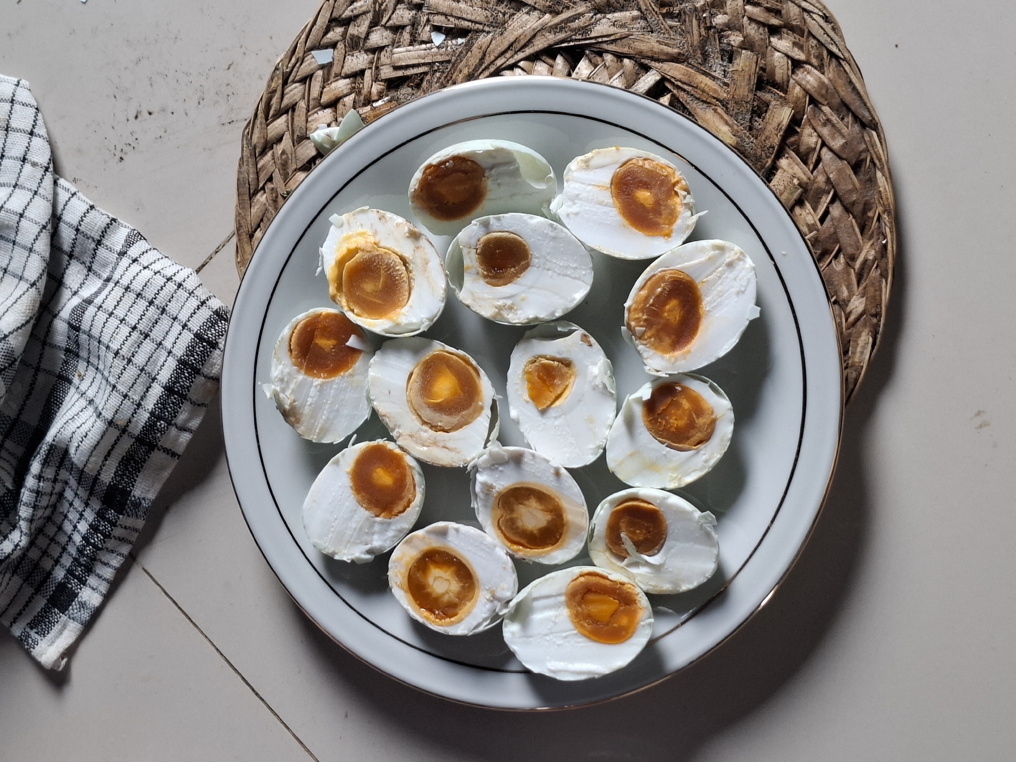 In Deah Pangwa, Indonesia, a white plate with a gold rim sits on a woven mat. The plate holds approximately twelve halves of salted duck eggs, revealing their distinct white albumen and vibrant orange yolks. A black and white checkered cloth lies draped across the tiled floor in the lower right corner of the image.