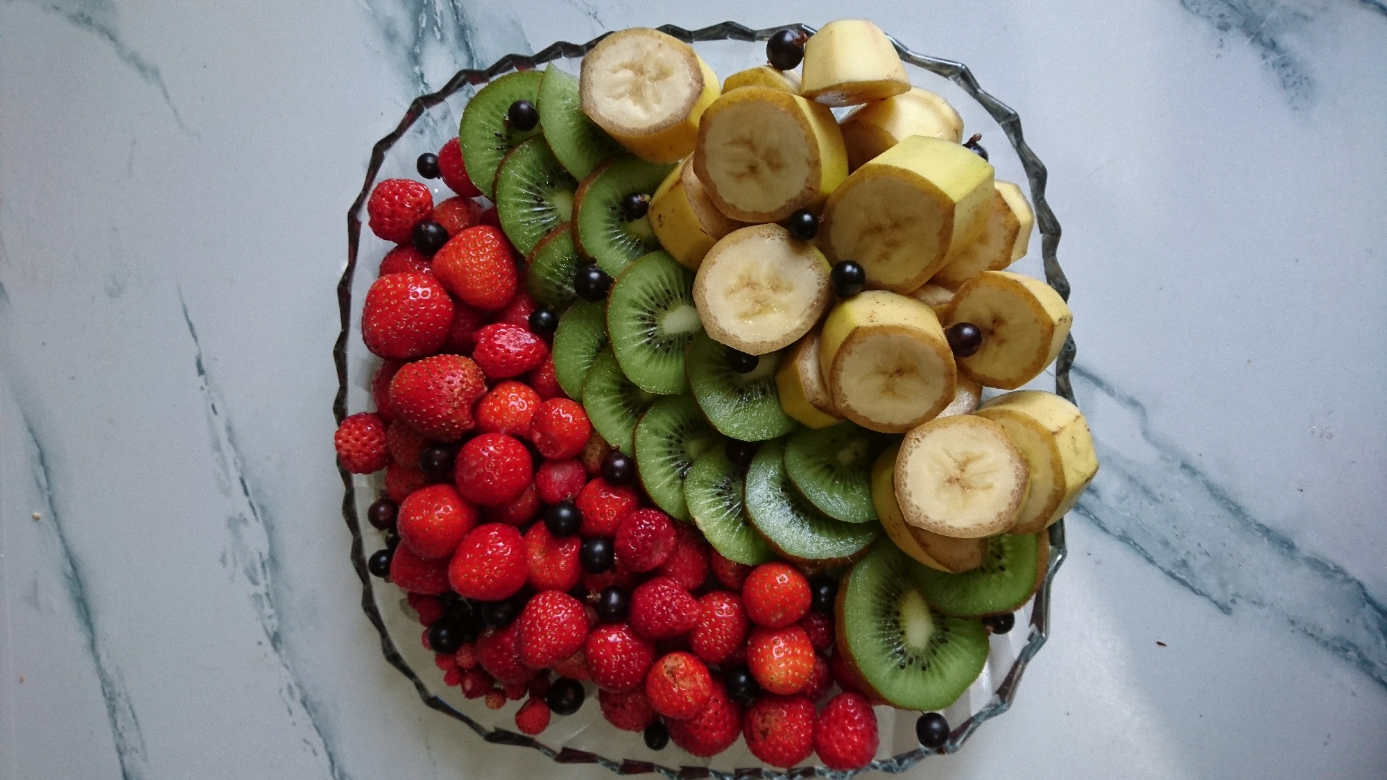 A clear glass platter, with a decorative, rippled edge, is filled with a colorful assortment of fruits, placed on a light-colored marble countertop. The platter is arranged with three distinct sections. On the left side, numerous small, red strawberries, some whole and some halved, are interspersed with dark, small berries, likely currants or blueberries. In the middle section, several circular slices of kiwi fruit are arranged in a slightly overlapping fan pattern, revealing their characteristic green flesh with dark seeds. To the right, slices of banana, cut into rounds, are stacked and fanned out. Some of the banana slices show a slightly browned edge. Small, dark berries are also scattered among the banana slices. The overall scene is well-lit, highlighting the fresh appearance of the fruit. There are no people visible in the image, and the setting does not provide any specific information that would suggest a location in Dvirets, Ukraine.