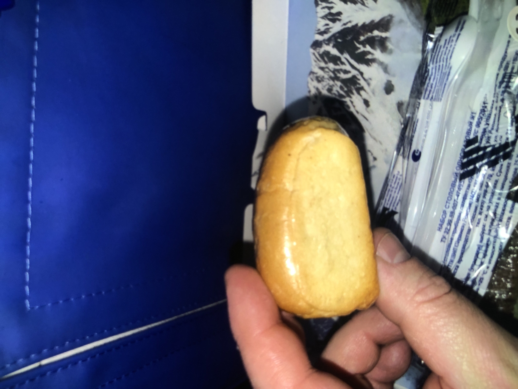 The image shows a person's hand holding a small, golden-brown, elongated bun. To the left of the bun is a blue object with white stitching, possibly fabric or a bag. To the right of the bun is a package of disposable utensils. Russian text is visible on the package. The bun is the primary focus of the image, held against the backdrop of the blue object and the packaged utensils.