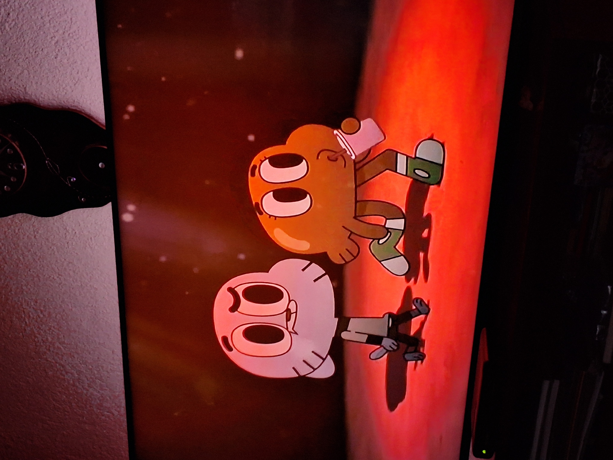 The image shows a screen displaying a cartoon featuring two anthropomorphic characters. One character, resembling a light-colored cotton ball with visible facial features, is positioned below a second character that resembles an orange peel with similar features. Both characters appear to be standing. The background is a gradient of red tones. The screen is positioned near a wall with a decorative object. The location is not discernible from the image. There is no evidence that it is located in Panagyurishte, Bulgaria.