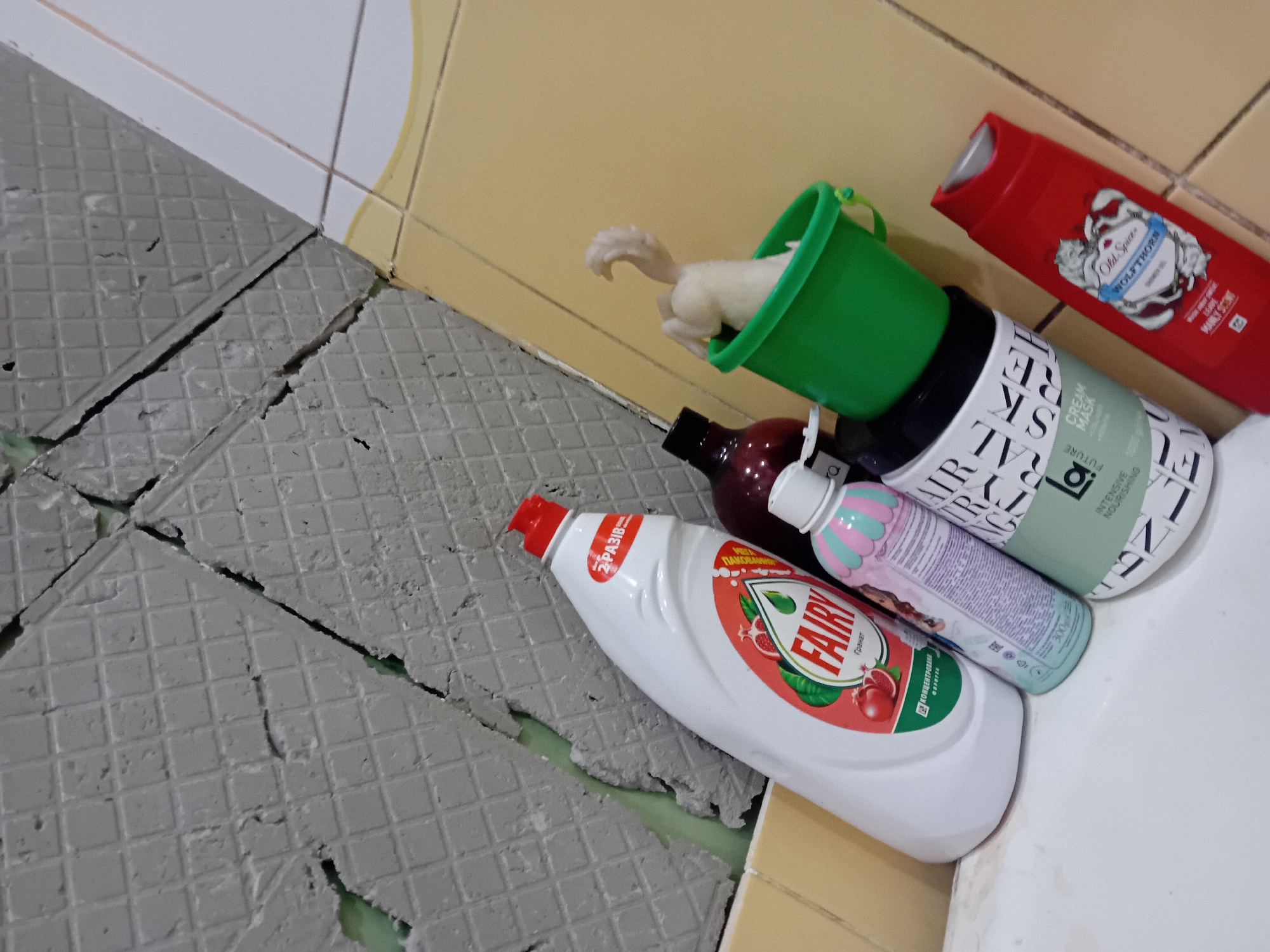 In Chuhuiv, Ukraine, a bathroom scene unfolds with items on a bathtub ledge against a backdrop of damaged, gray square tiles and yellow tiles. Dominating the foreground is a large white bottle of "Fairy" dish soap with a red cap and a pomegranate graphic. To its right, a small, pastel pink and blue bottle stands, followed by a dark bottle with a black cap. Further right, a green bucket holds a light-colored, abstract object resembling a toy or decoration. To the right of the bucket, a white bottle with black lettering and a green label is positioned. Finally, a red bottle of "Old Spice" body wash with a wolf graphic rests on the far right. The tiles above the bathtub appear to be in disrepair, with visible gaps and exposed adhesive.