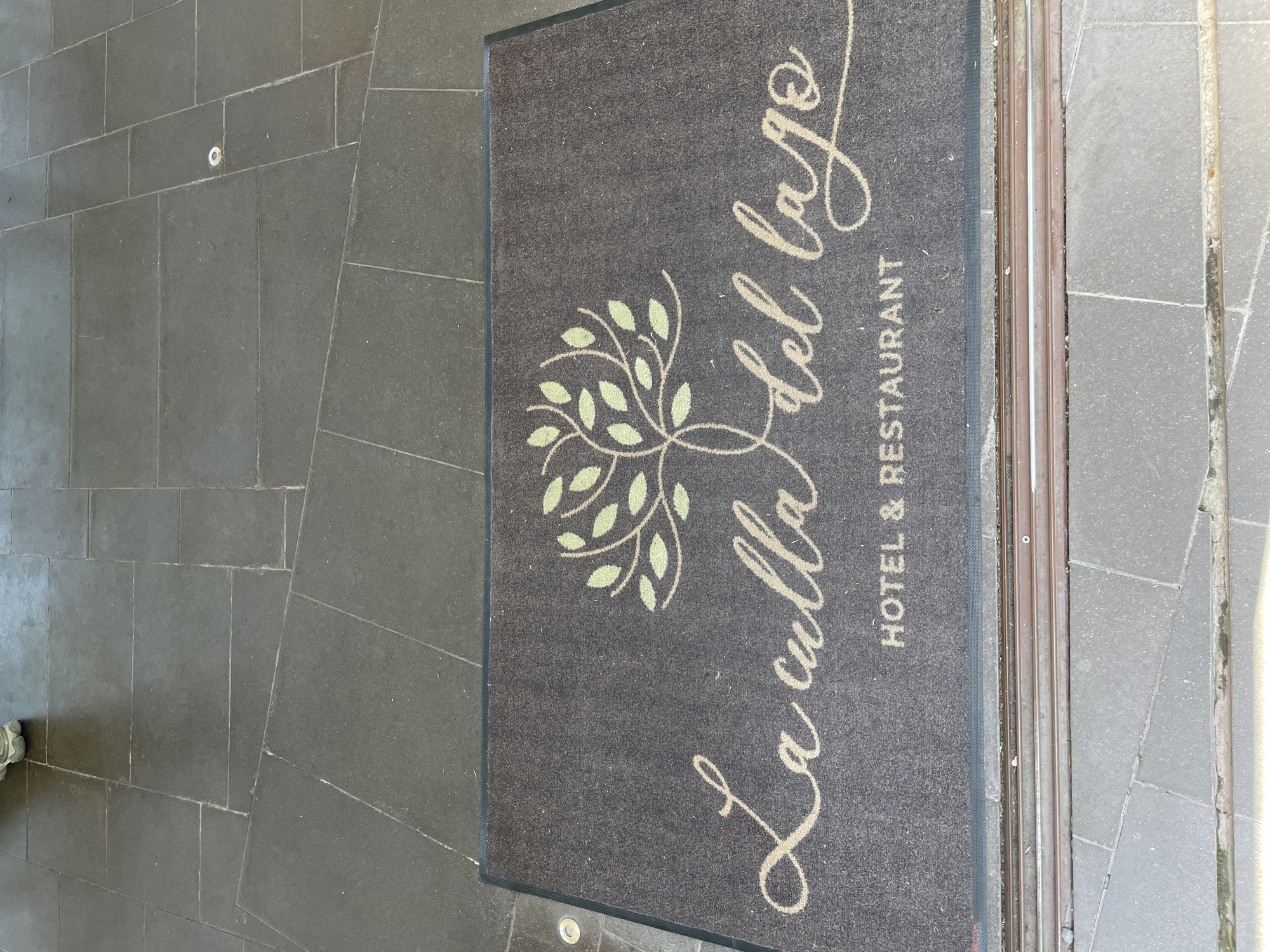 The image shows a doormat outside a building. The doormat is grey with a green logo of a tree and the name "La Culla del Lago" written in a cursive font. Below the name, it says "Hotel & Restaurant". The doormat is lying on a stone floor, and part of a doorway is visible on the right. The doormat is located in Castel Gandolfo, Italy.